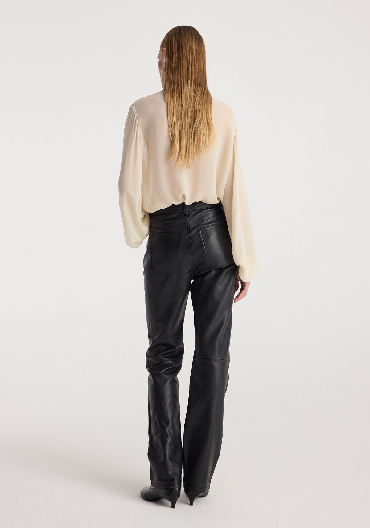 Woman wearing a cream-colored Romantic top | cream with long sleeves, paired with high-waisted black leather pants, standing facing away against a white background.