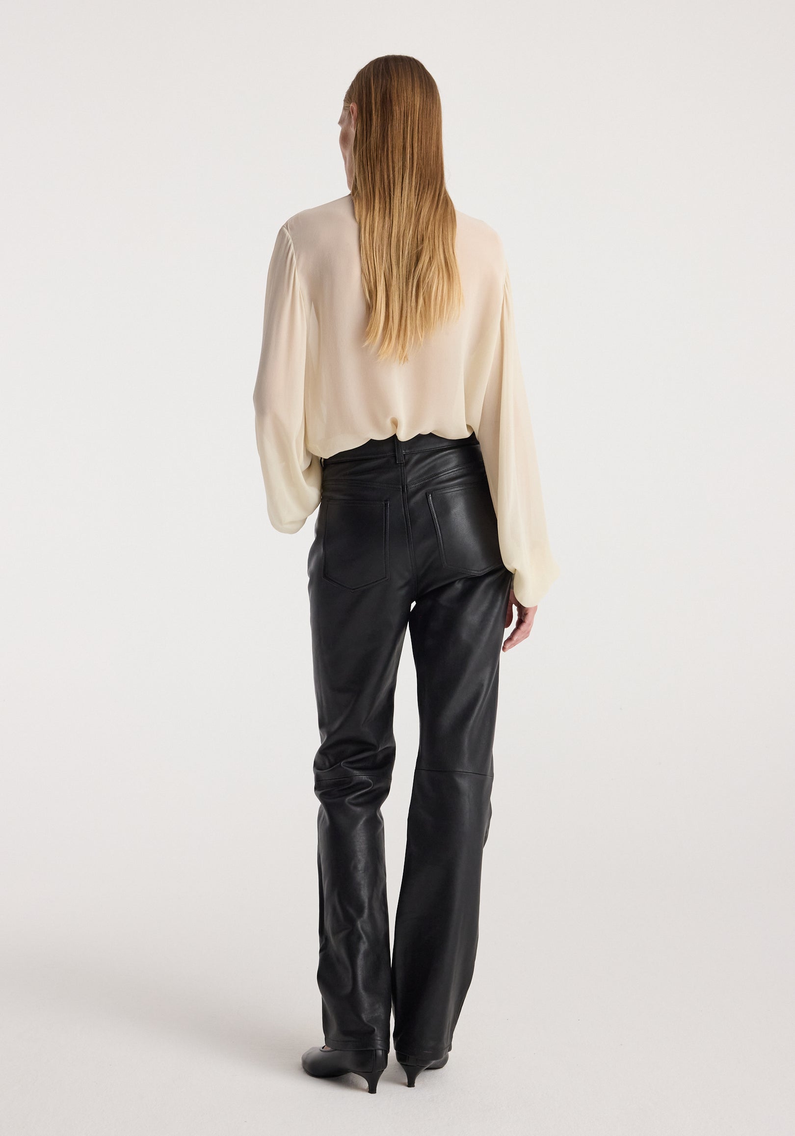 Woman wearing a cream-colored Romantic top | cream with long sleeves, paired with high-waisted black leather pants, standing facing away against a white background.