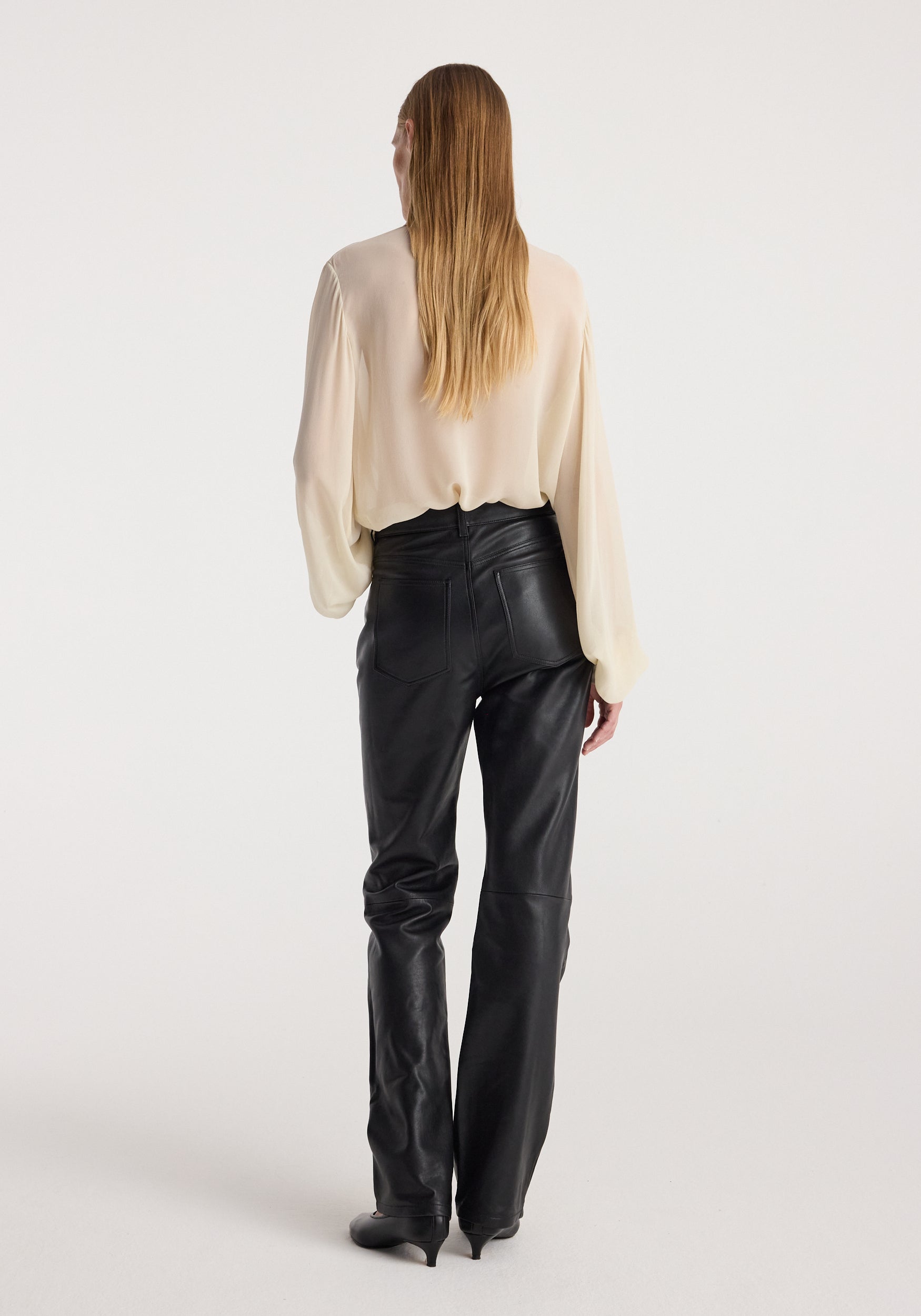 Woman wearing a cream-colored Romantic top | cream with long sleeves, paired with high-waisted black leather pants, standing facing away against a white background.
