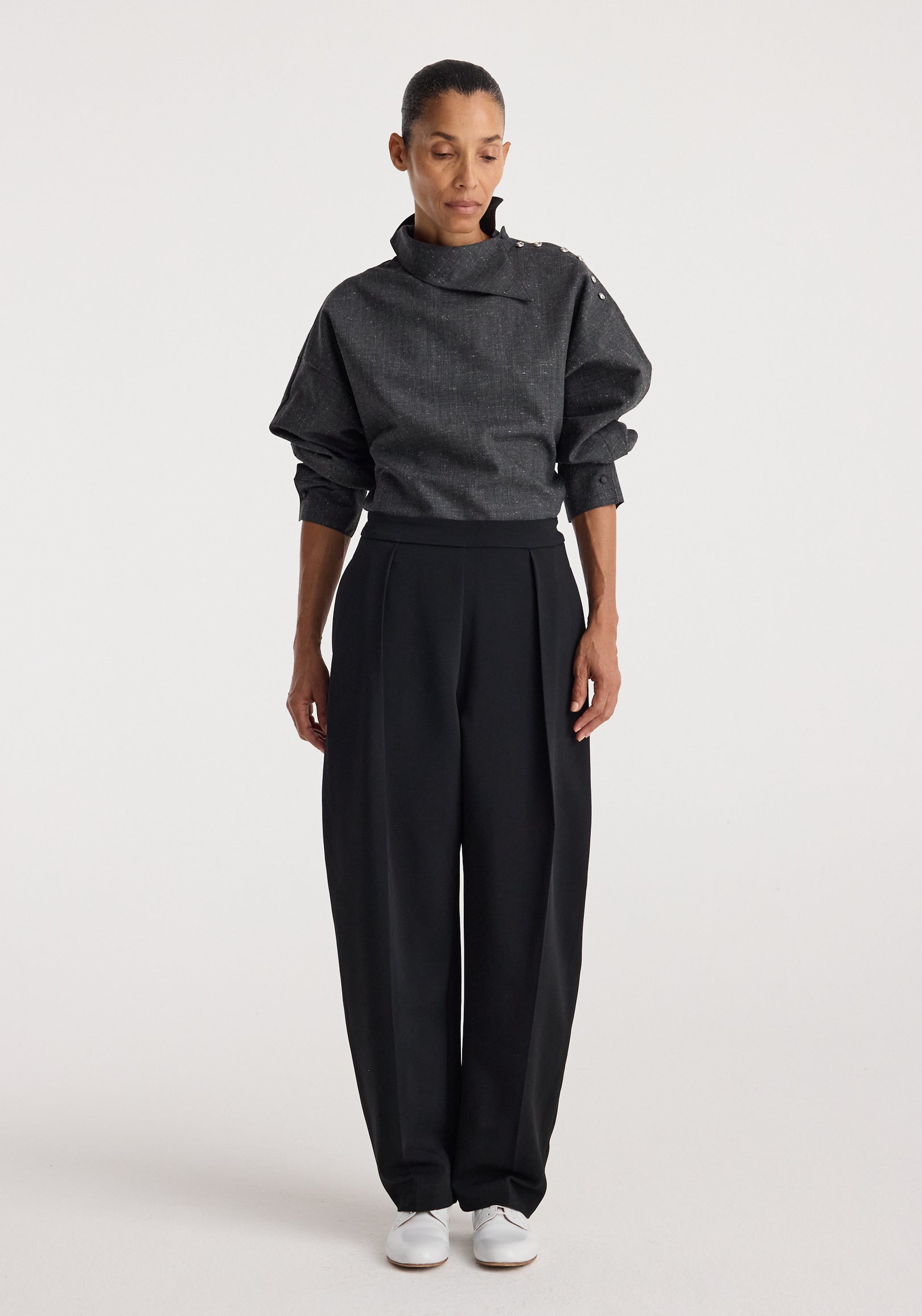 Woman wearing a grey slub fabric top with a high, structured collar and button details on the shoulder, styled with loose black trousers and white shoes, Reimagined collar top | grey slub.