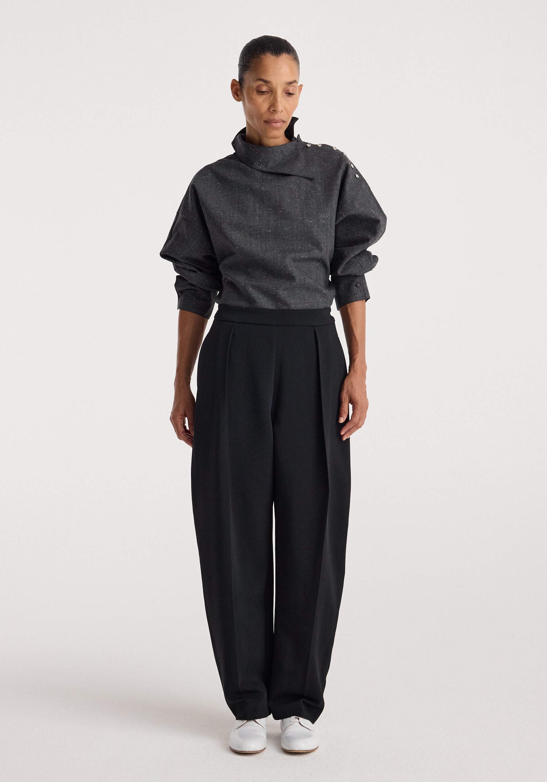Woman wearing a grey slub fabric top with a high, structured collar and button details on the shoulder, styled with loose black trousers and white shoes, Reimagined collar top | grey slub.