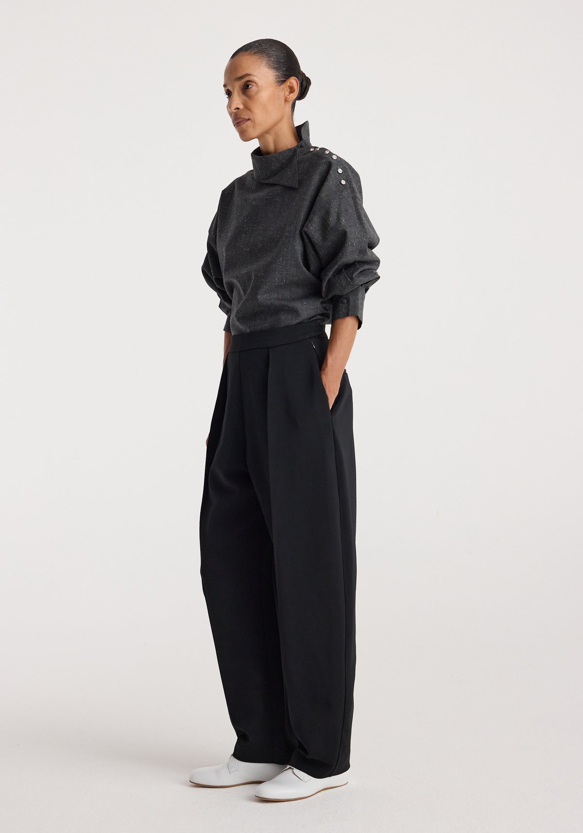 Woman wearing a grey slub textured Reimagined collar top | grey slub with long sleeves featuring shoulder buttons, paired with loose black trousers and white shoes.