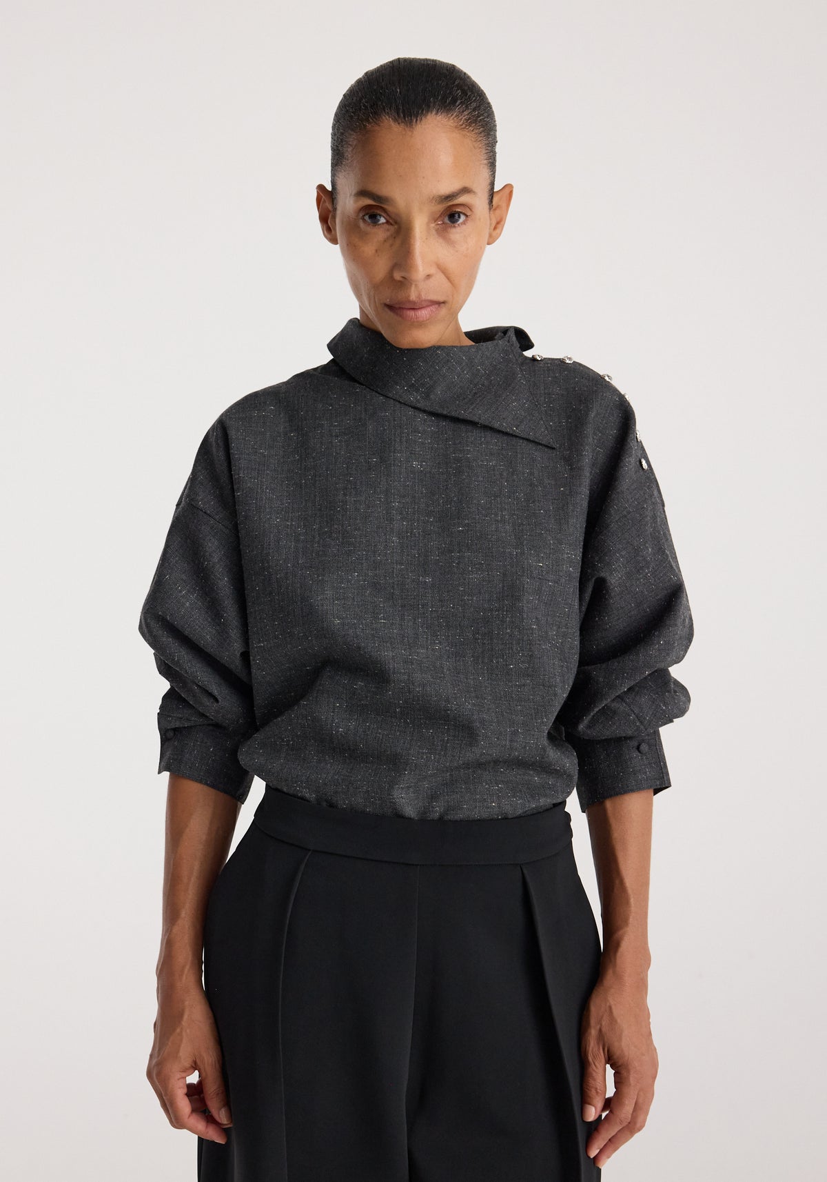 Woman wearing a dark grey textured long-sleeve top with a structured, asymmetrical collar and button detail, styled with black high-waisted pants, Reimagined collar top | grey slub