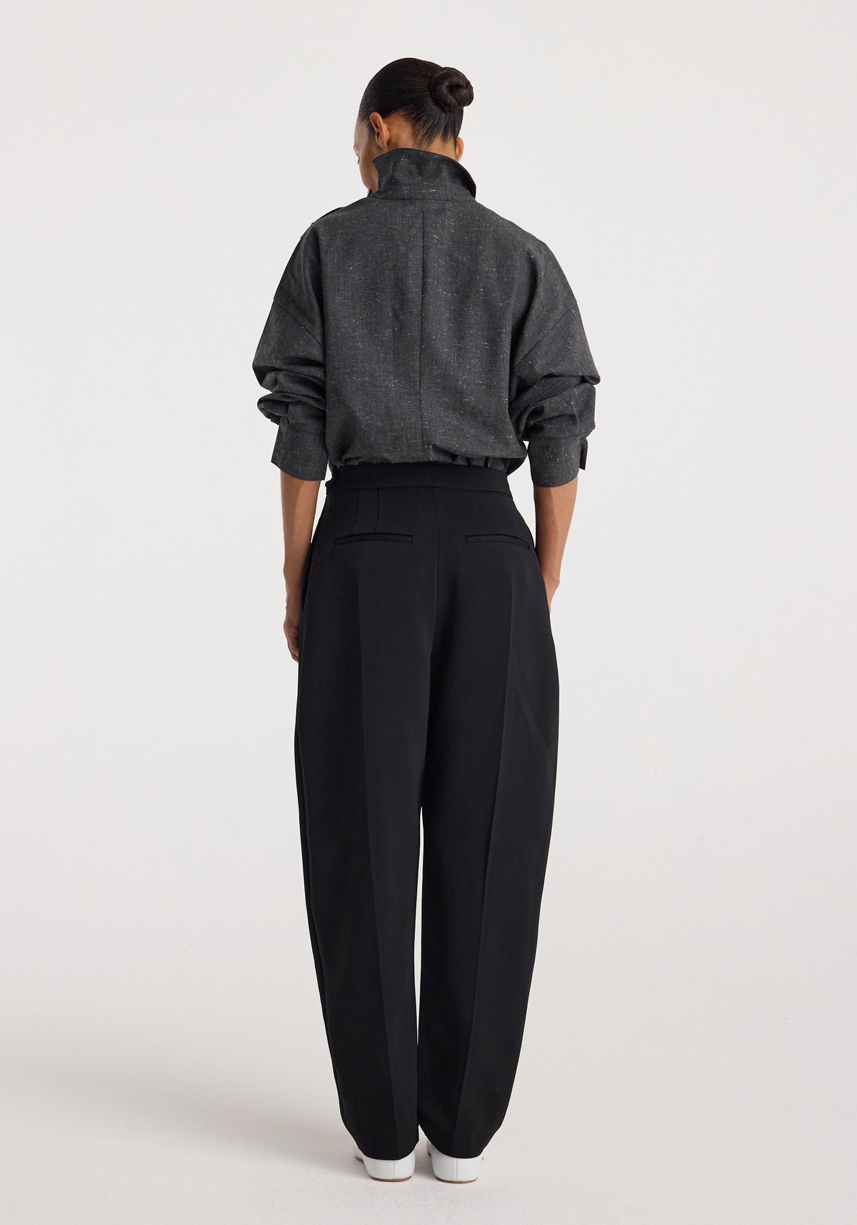Woman wearing a dark grey slub textured "Reimagined collar top | grey slub" with long sleeves, standing facing away, paired with high-waisted black trousers and white shoes.