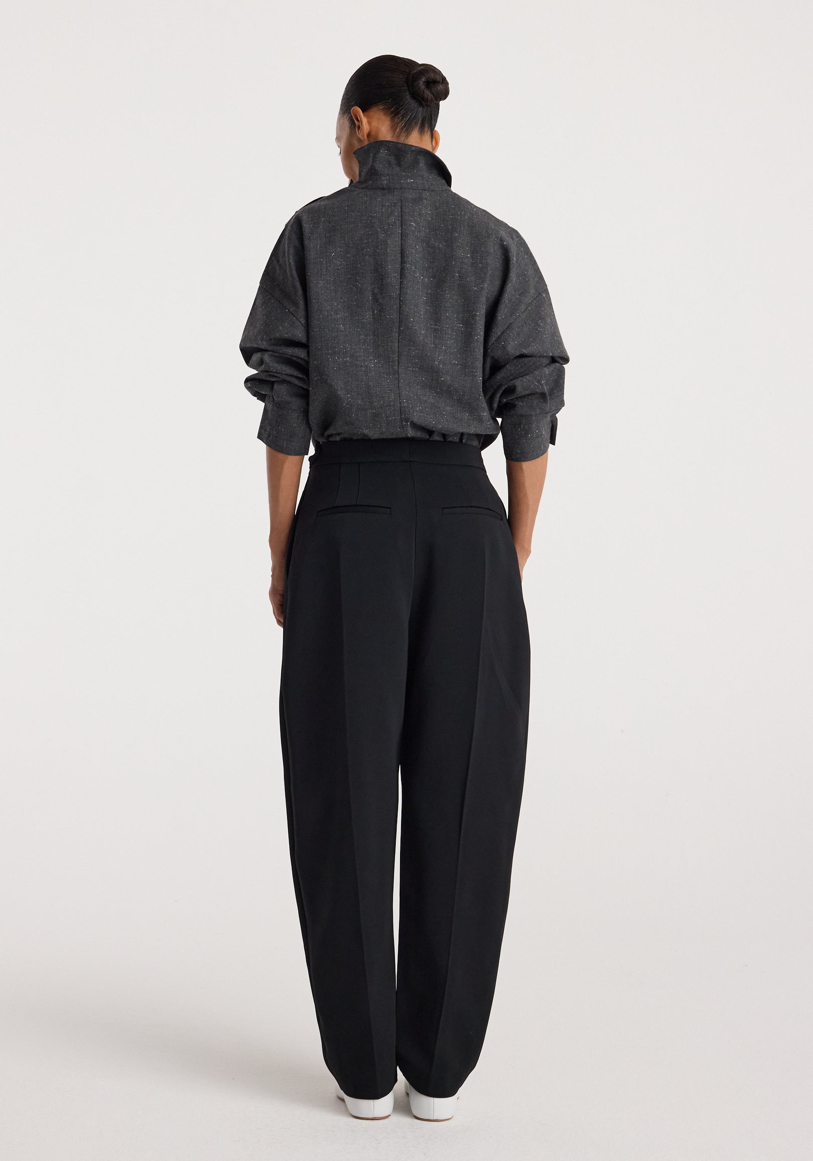 Woman wearing a dark grey slub textured "Reimagined collar top | grey slub" with long sleeves, standing facing away, paired with high-waisted black trousers and white shoes.