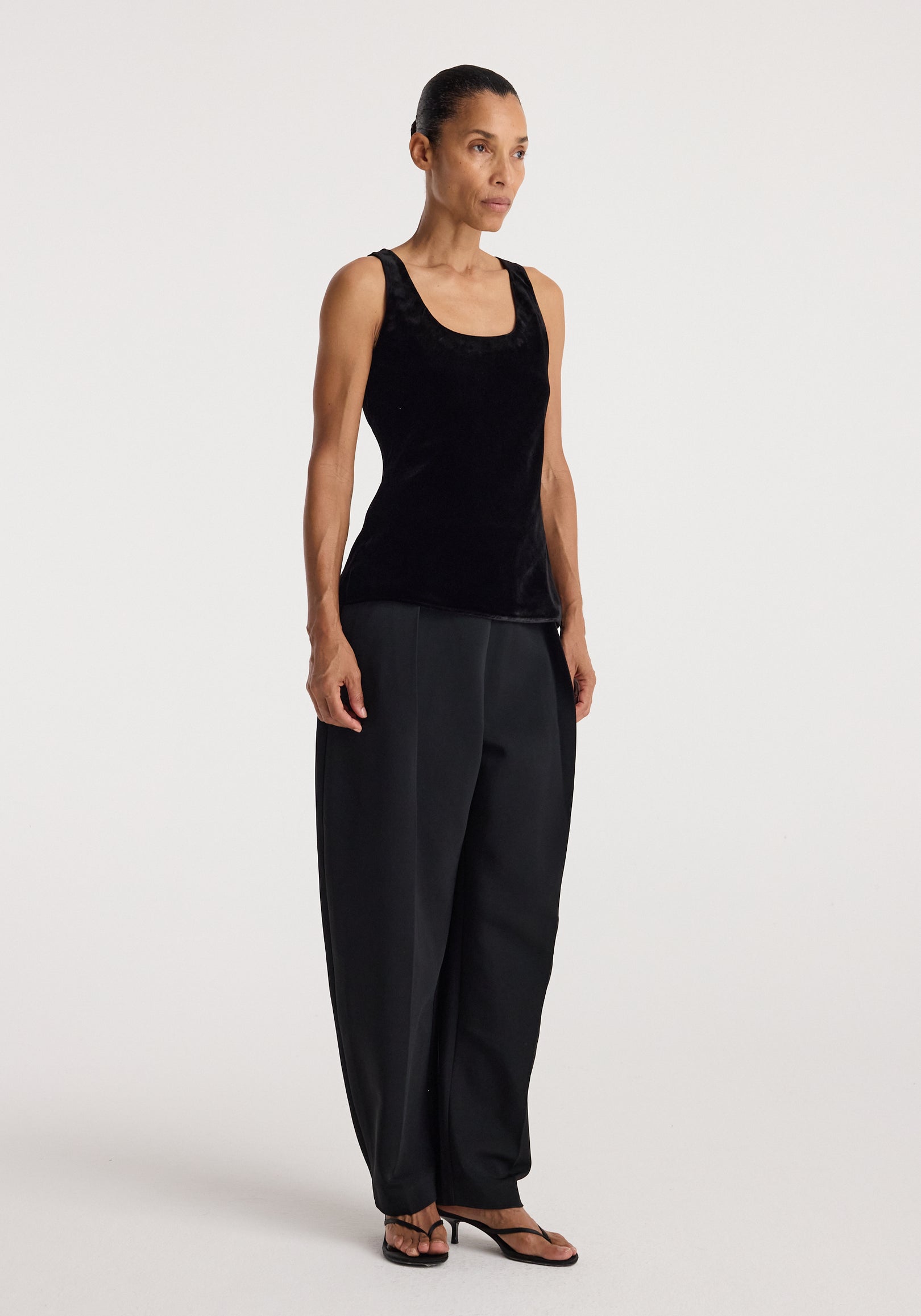 Woman wearing a black Scoop-neck silk velvet top | black paired with loose black trousers and open-toe heels, standing against a plain light background.