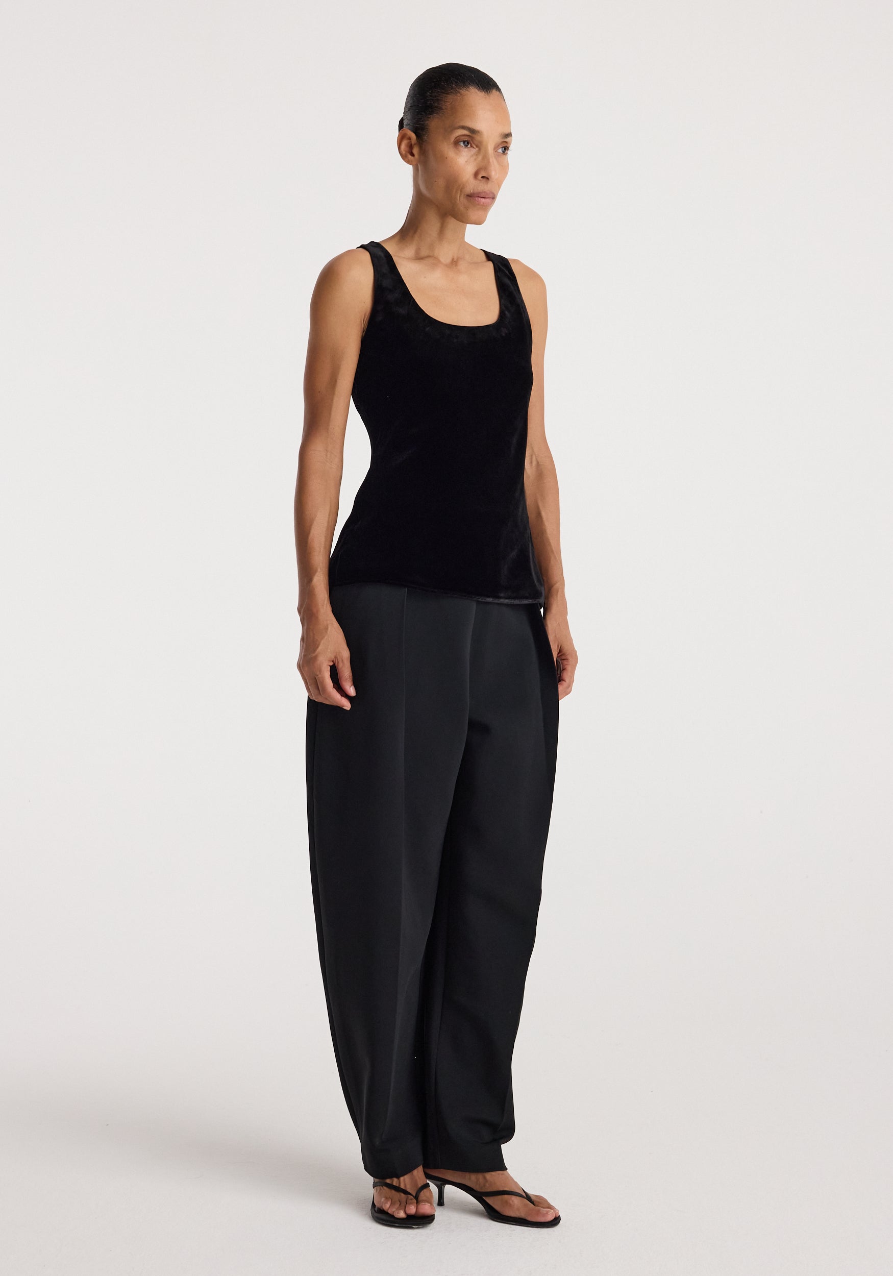 Woman wearing a black Scoop-neck silk velvet top | black paired with loose black trousers and open-toe heels, standing against a plain light background.