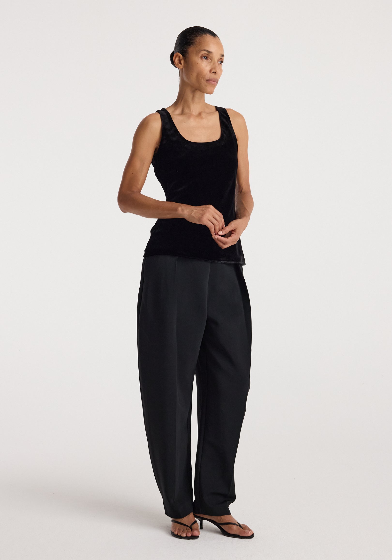 Woman wearing a black scoop-neck silk velvet top | black paired with loose black pants and black open-toe sandals, standing against a plain white background.