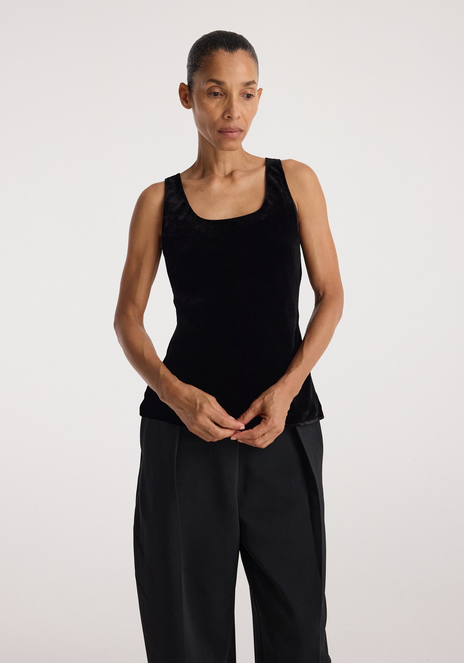 Woman wearing a black scoop-neck silk velvet top and loose black pants standing against a plain background with her hands clasped in front of her.