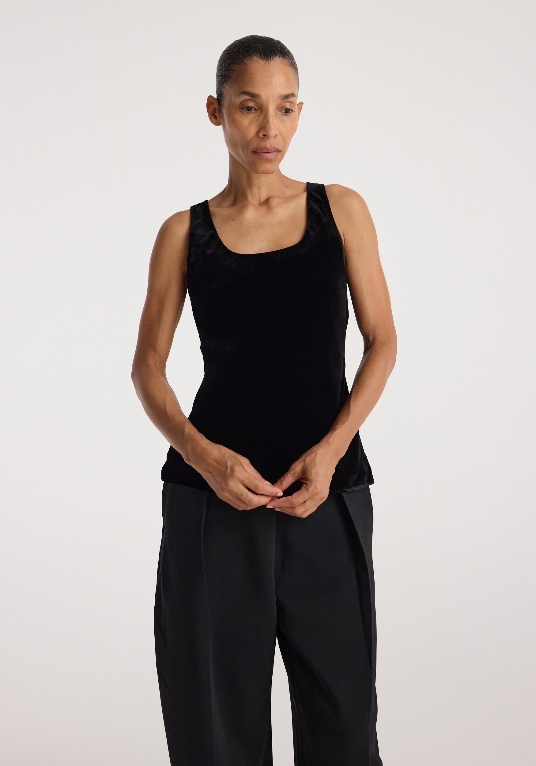 Woman wearing a black scoop-neck silk velvet top and loose black pants standing against a plain background with her hands clasped in front of her.