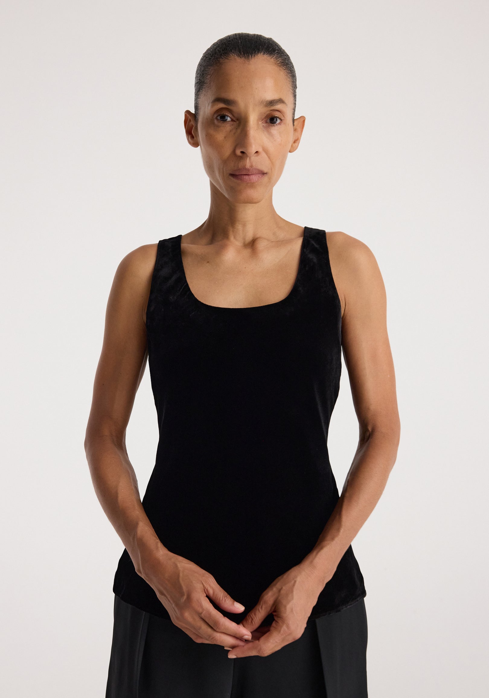 Woman wearing a sleeveless, fitted Scoop-neck silk velvet top | black, standing against a plain background with hands loosely clasped in front.