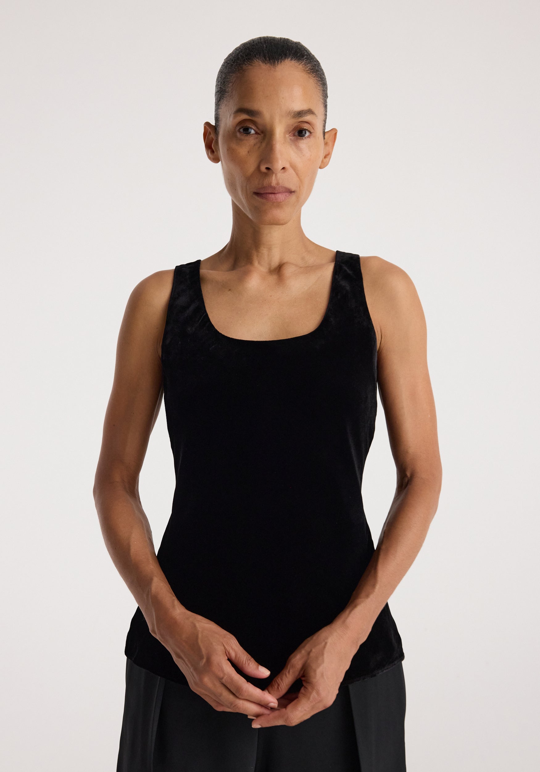 Woman wearing a sleeveless, fitted Scoop-neck silk velvet top | black, standing against a plain background with hands loosely clasped in front.