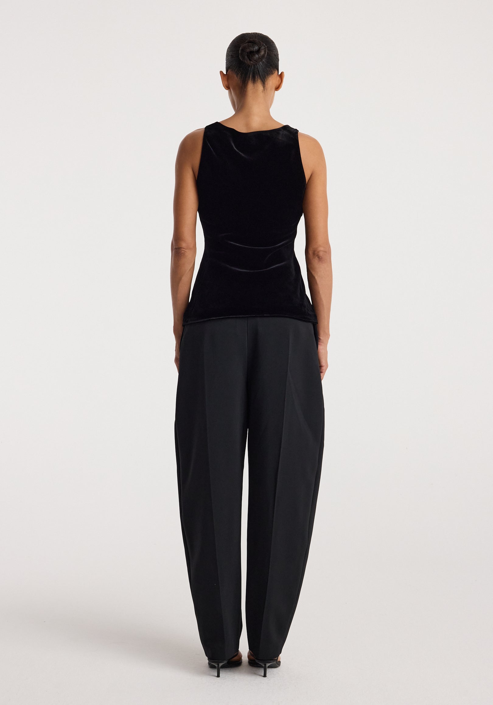 Woman seen from behind wearing a black scoop-neck silk velvet top | black paired with loose black trousers and black heels against a plain background.