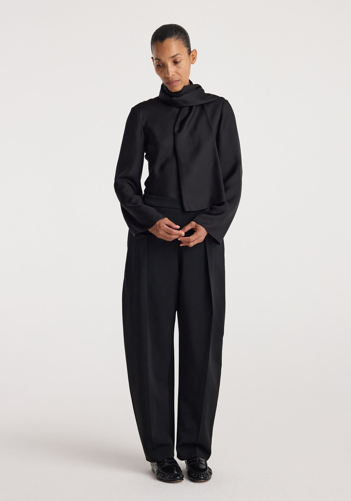 Woman wearing a black silk foulard top | black paired with matching wide-leg trousers and black shoes standing against a plain white background.