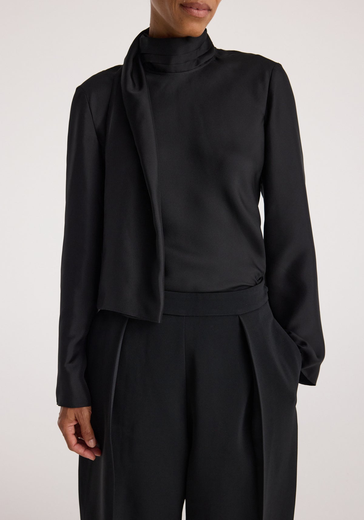 Woman wearing a sleek black long-sleeve Silk foulard top | black with a draped detail over the shoulder, paired with matching high-waisted black trousers.