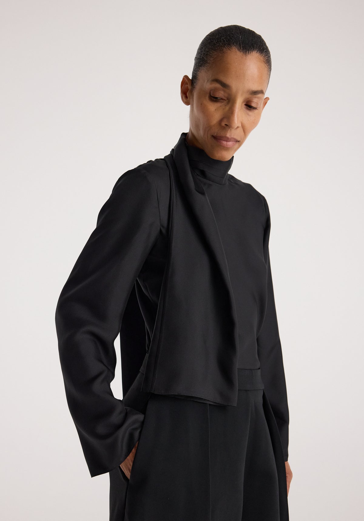 Woman wearing a sophisticated black Silk foulard top | black with long sleeves and a high neckline, styled with matching black pants against a neutral background.
