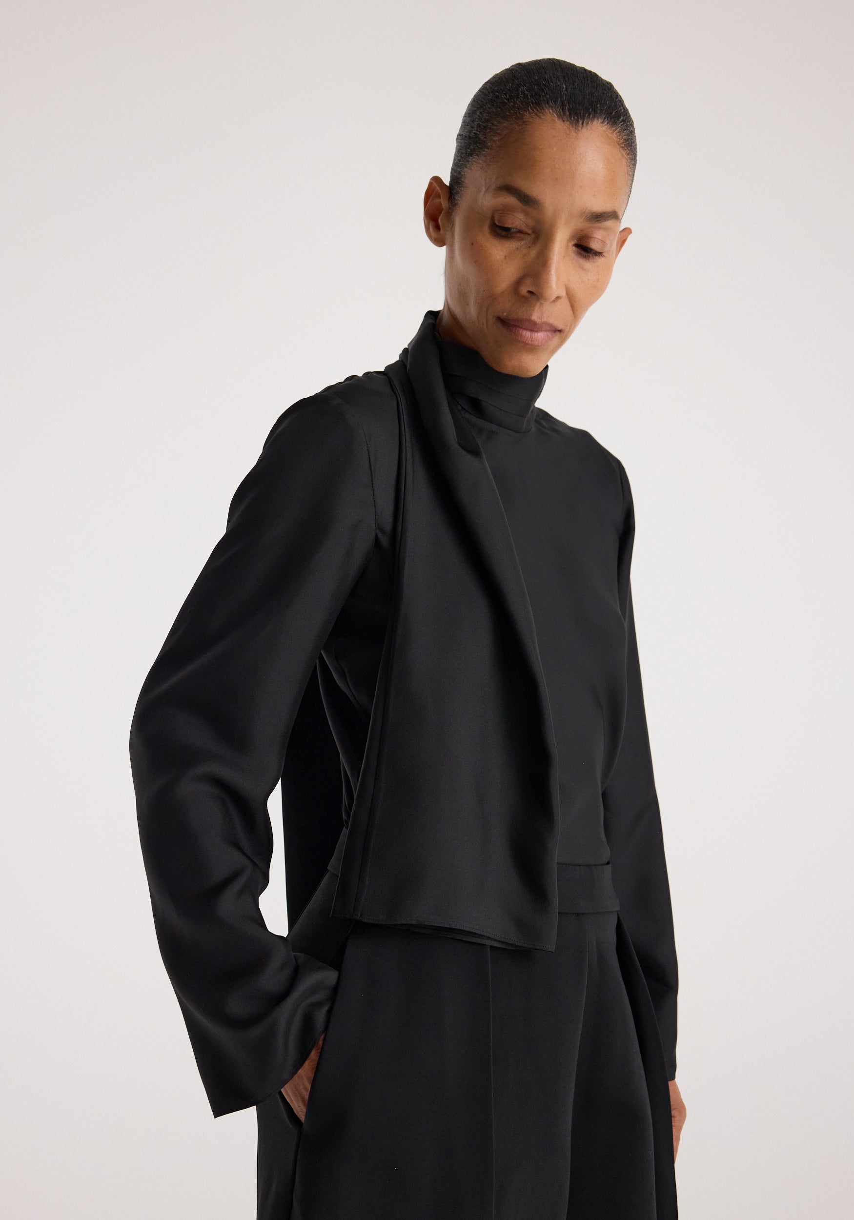 Woman wearing a sophisticated black Silk foulard top | black with long sleeves and a high neckline, styled with matching black pants against a neutral background.