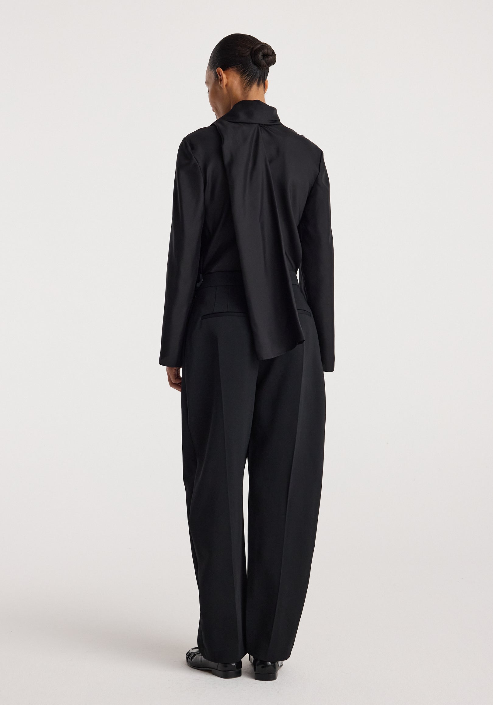 Back view of a woman wearing a sleek black Silk foulard top | black with long sleeves paired with loose black trousers against a plain light background.