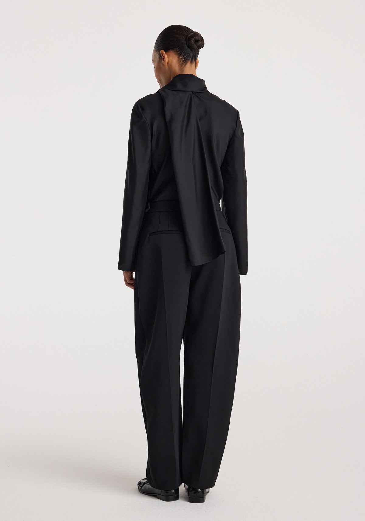 Back view of a woman wearing a sleek black Silk foulard top | black with long sleeves paired with loose black trousers against a plain light background.