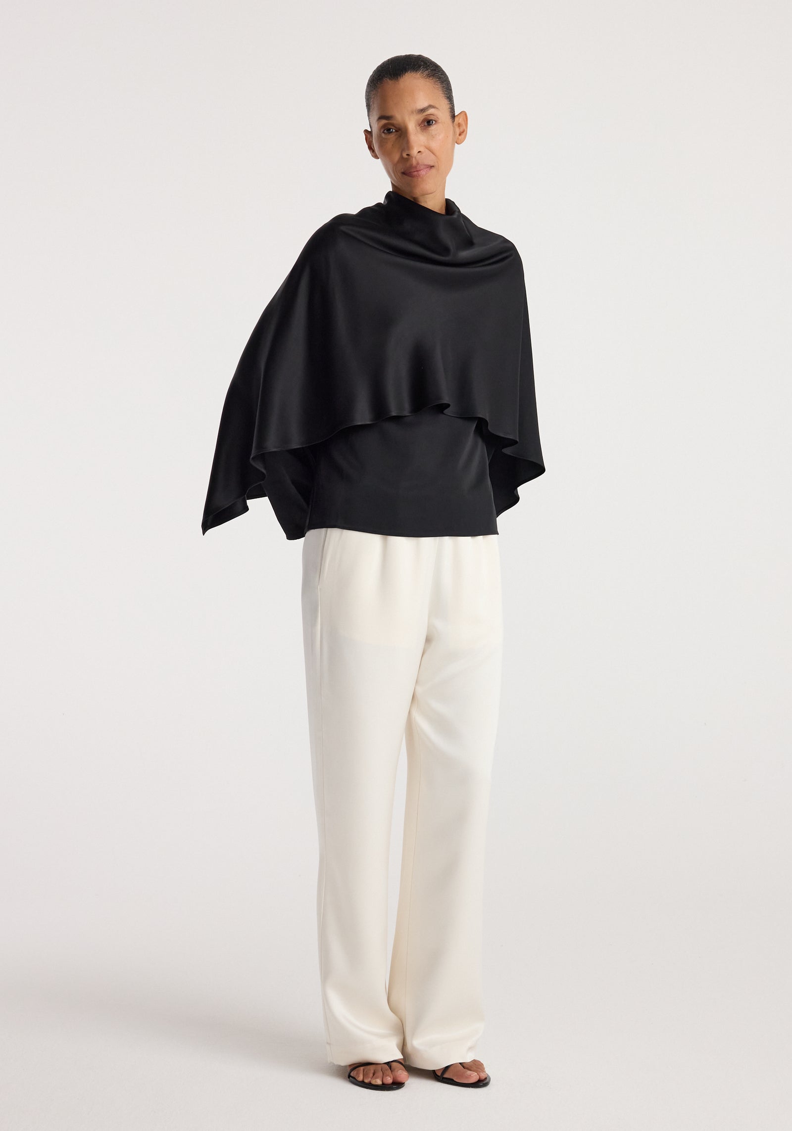 Woman wearing a black Satin cape top | black with a high neckline paired with loose white pants and black open-toe sandals, standing against a plain light background.