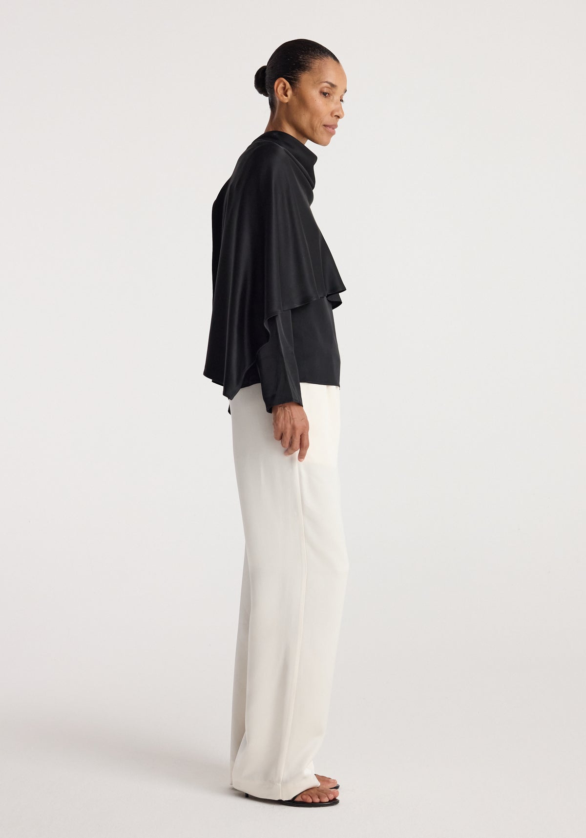 Woman wearing a black Satin cape top | black with draped sleeves paired with white wide-leg pants, standing in profile against a plain background.