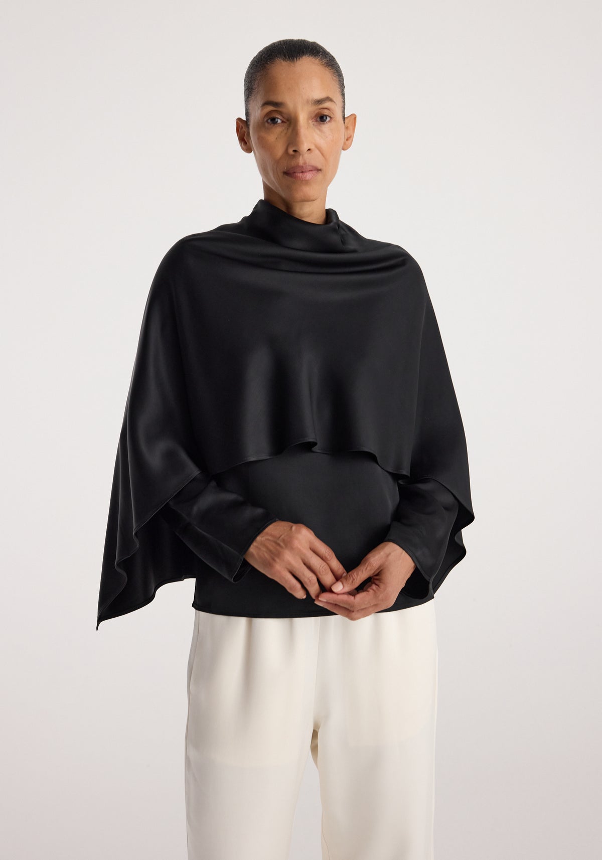 Woman wearing a black satin cape top | black with long sleeves and draped cape detail, paired with white high-waisted pants, standing against a plain background.