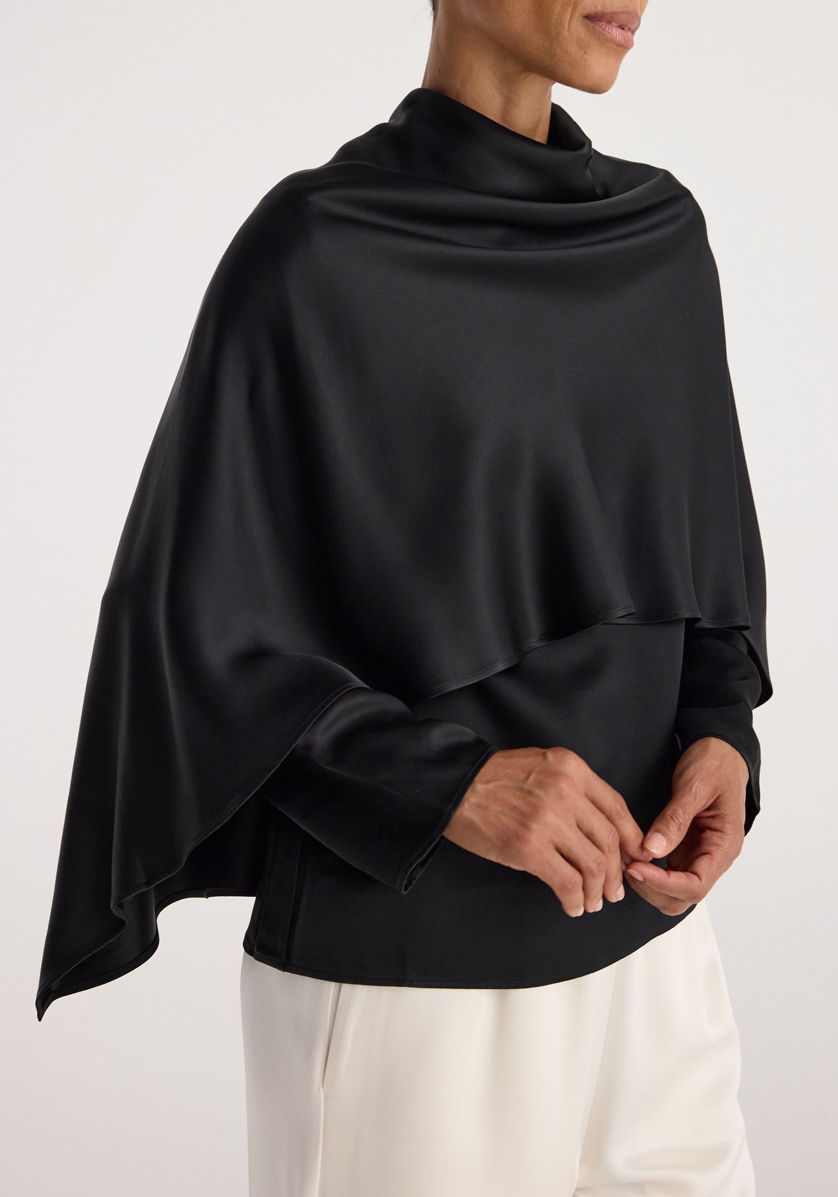 Woman wearing a smooth, flowing satin cape top | black with a high neckline and asymmetrical hem over a black long-sleeve shirt and white pants.