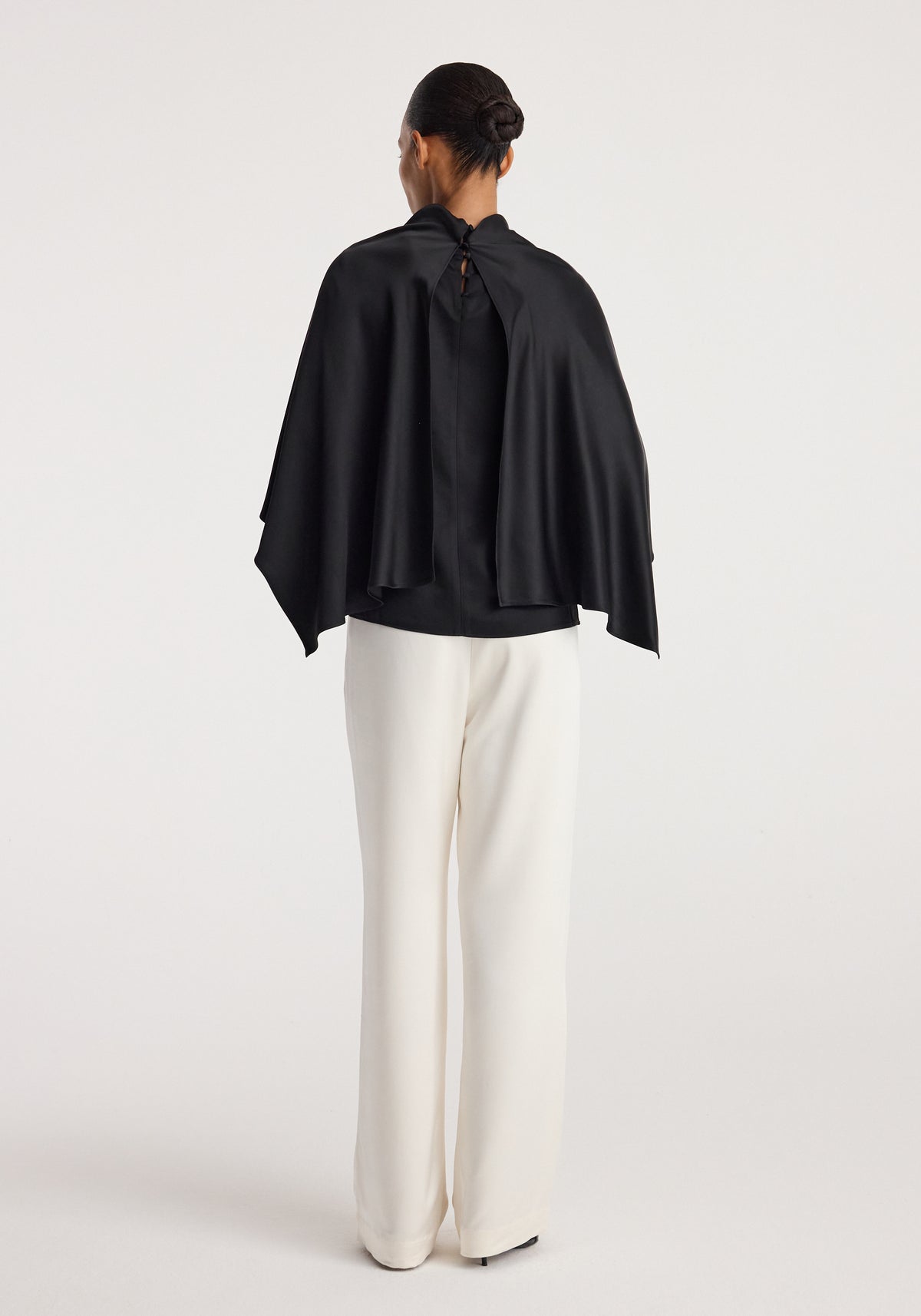 Woman wearing a black Satin cape top | black with a high neckline and flowing cape detail, paired with wide-leg white pants, standing against a plain background.