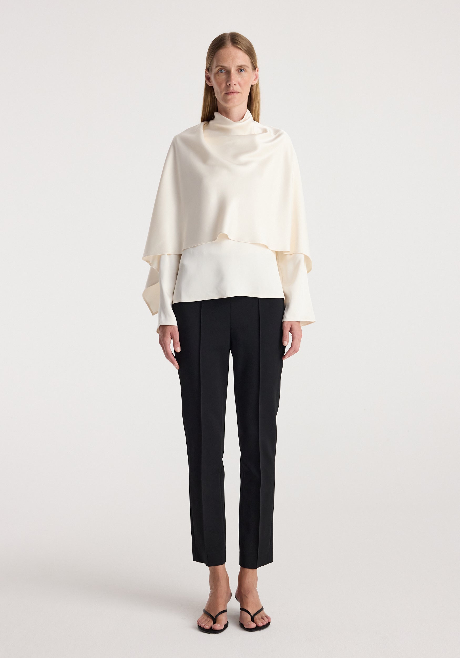 Model wearing a cream satin cape top | cream with long sleeves and a high neck, paired with black tailored pants and black open-toe sandals against a plain background.