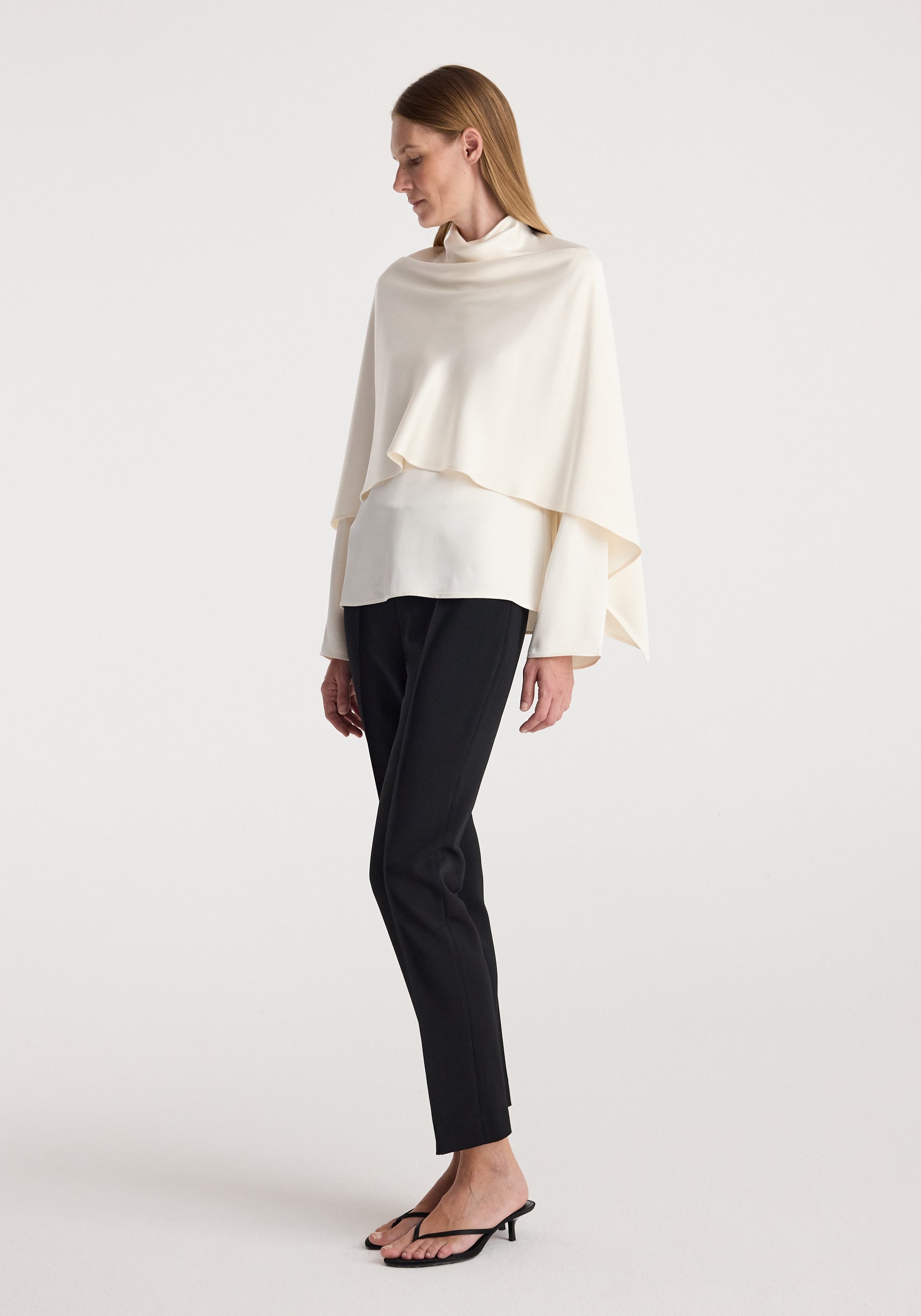 Woman wearing a cream satin cape top | cream with long sleeves, paired with black slim pants and black open-toe heels, standing against a plain white background.