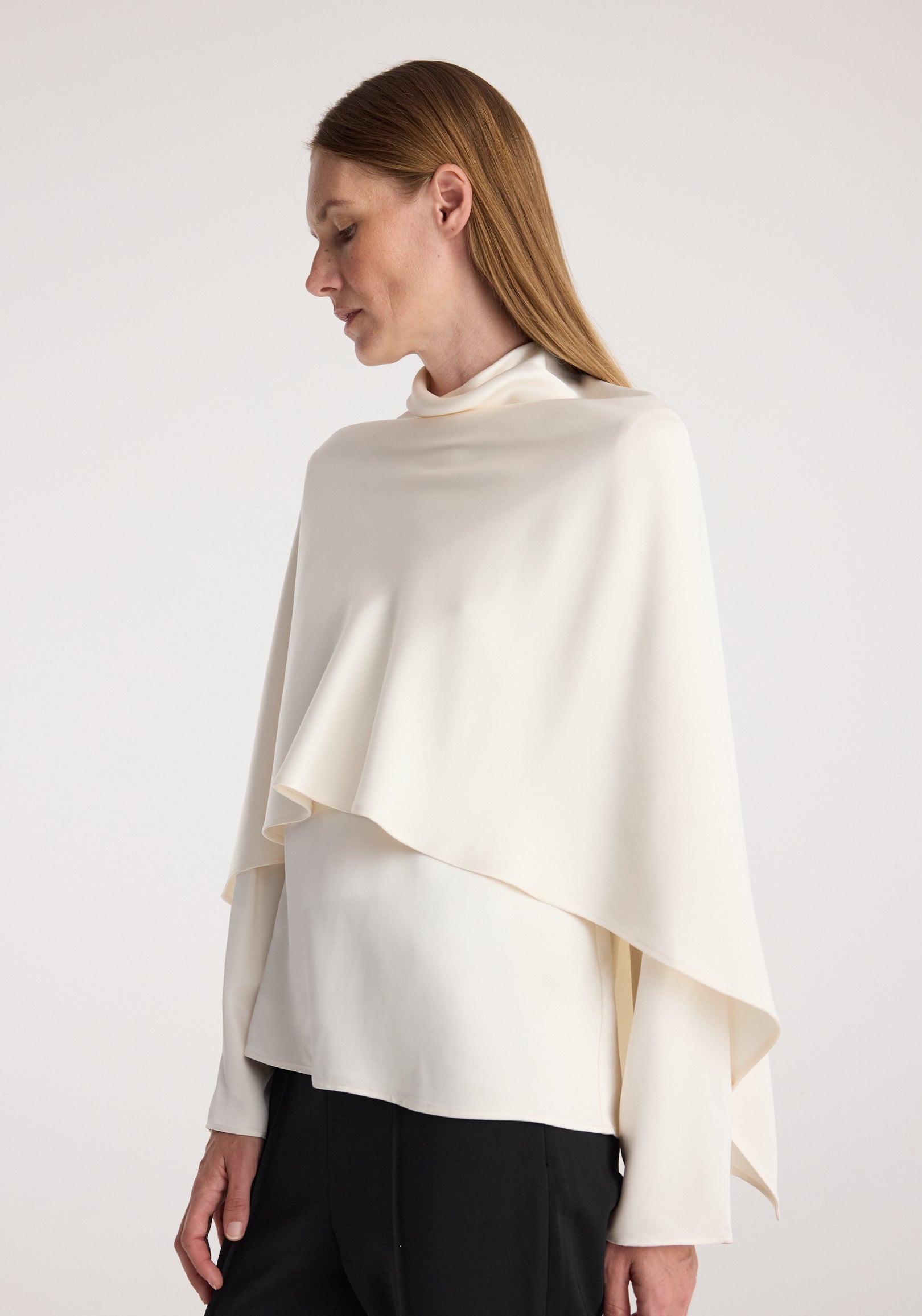 Woman wearing a cream satin cape top | cream with a high neckline and asymmetrical draped design, paired with black pants against a neutral background.