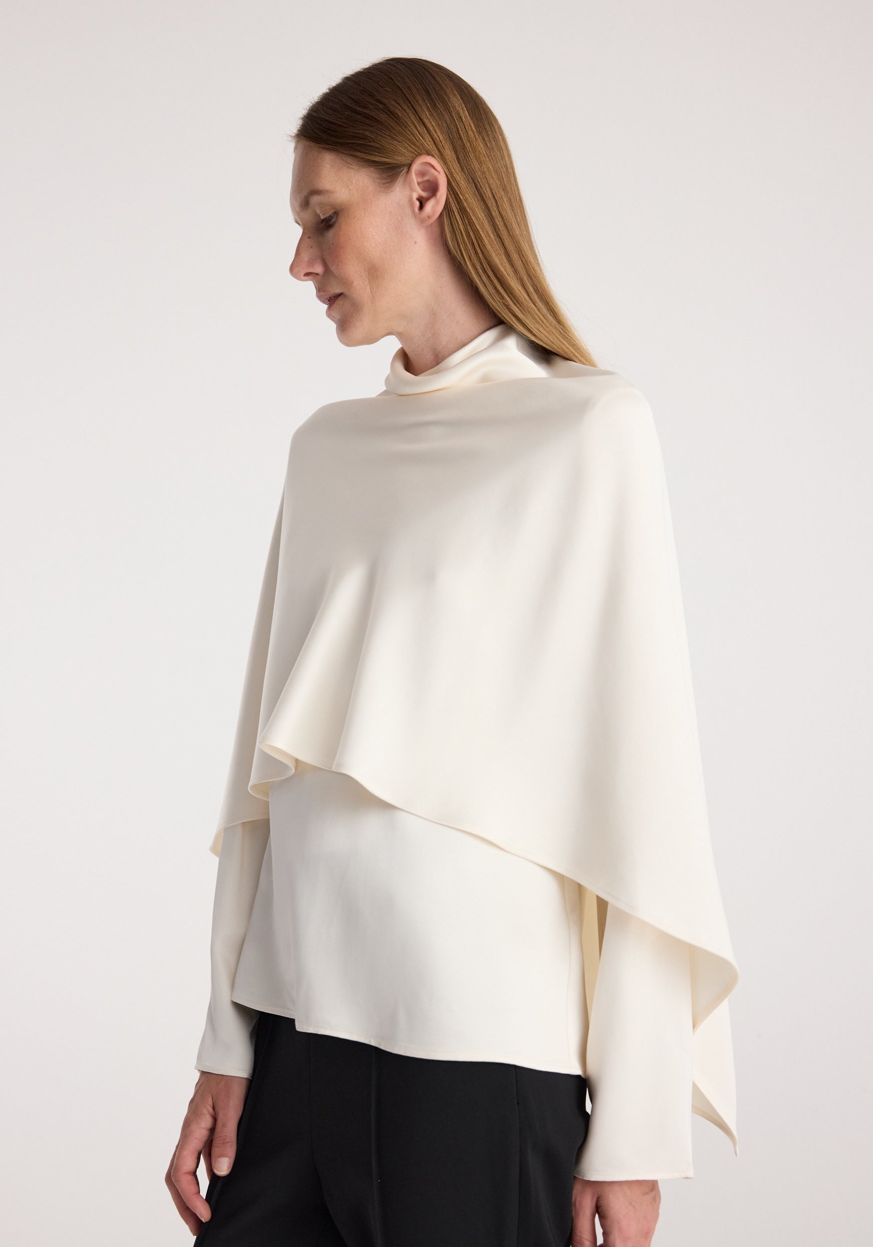 Woman wearing a cream satin cape top | cream with a high neckline and asymmetrical draped design, paired with black pants against a neutral background.