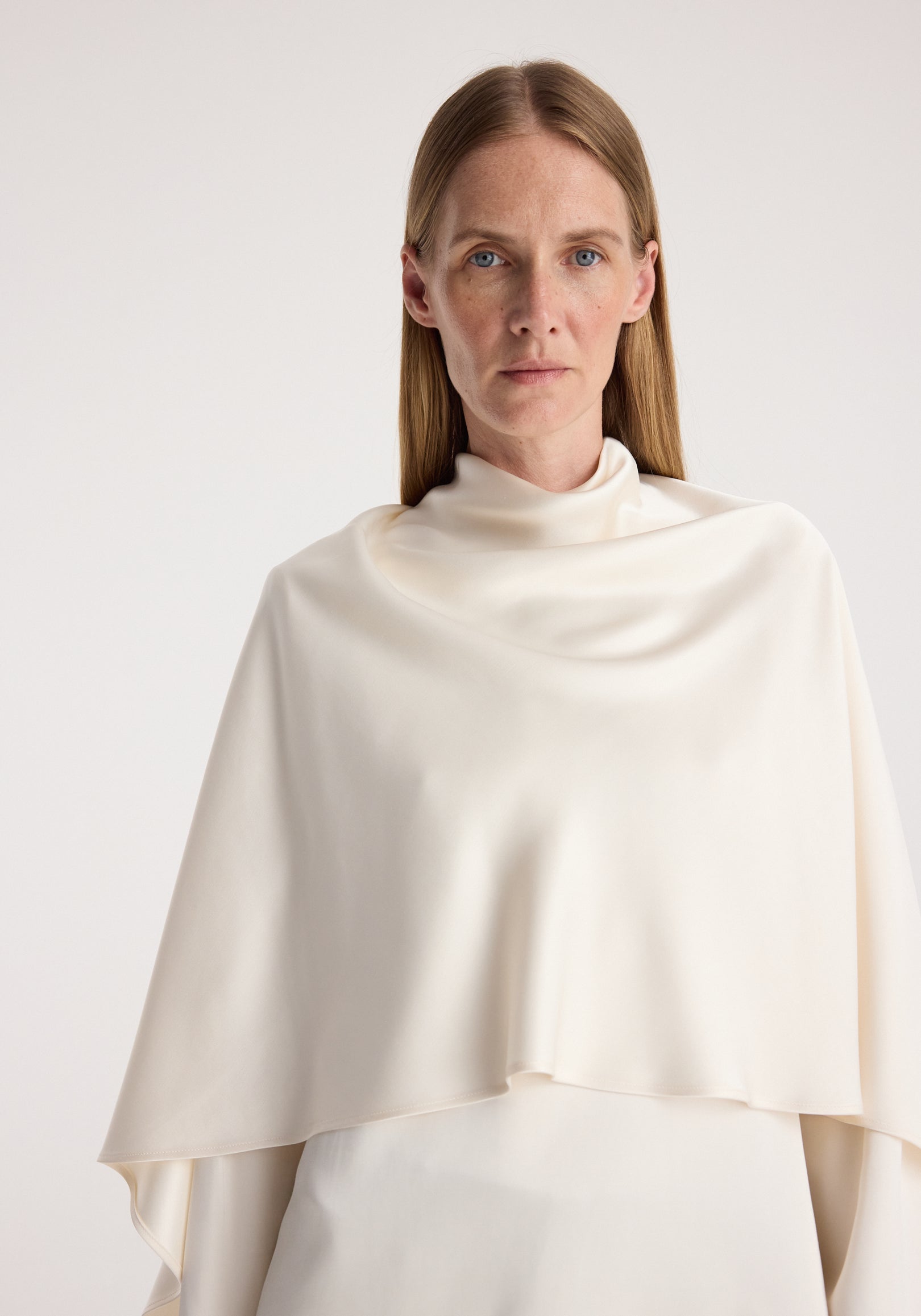 Woman wearing a cream satin cape top with a high neckline, showcasing smooth, flowing fabric and a minimalist design in a neutral studio setting.