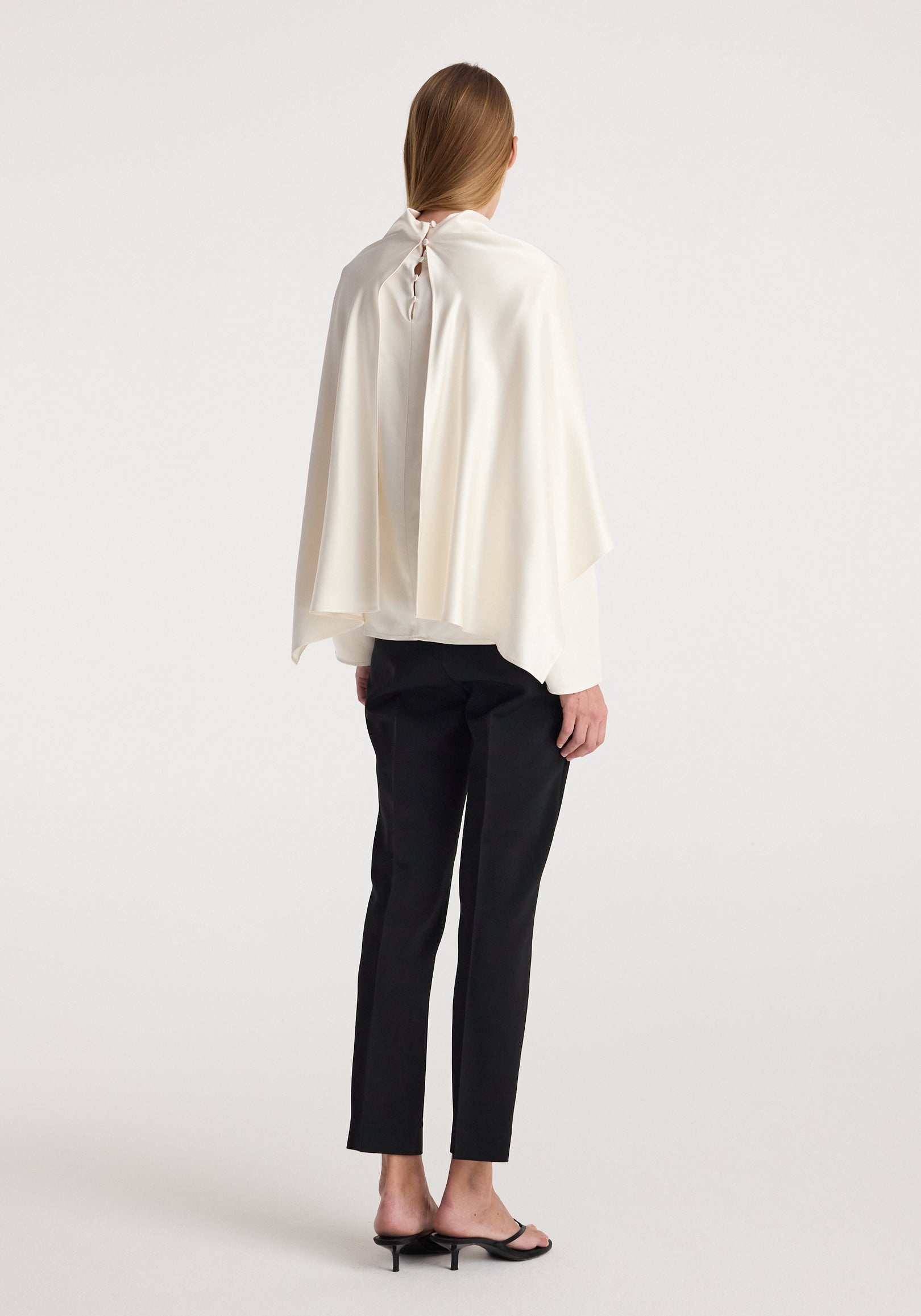 Woman seen from the back wearing a Satin cape top | cream with a loose, flowing fit and button closure at the neck, paired with black pants and black open-toe heels.