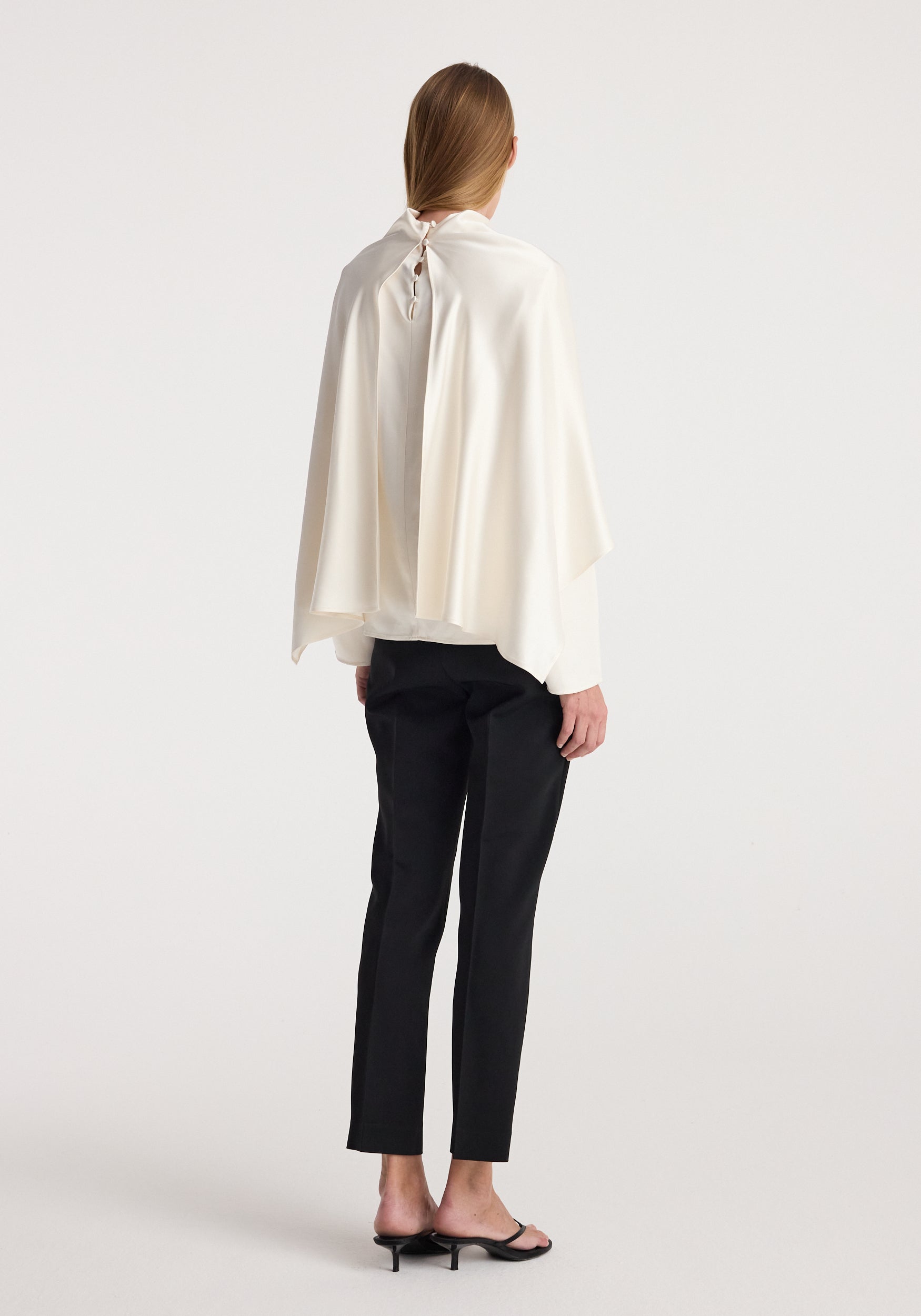 Woman seen from the back wearing a Satin cape top | cream with a loose, flowing fit and button closure at the neck, paired with black pants and black open-toe heels.