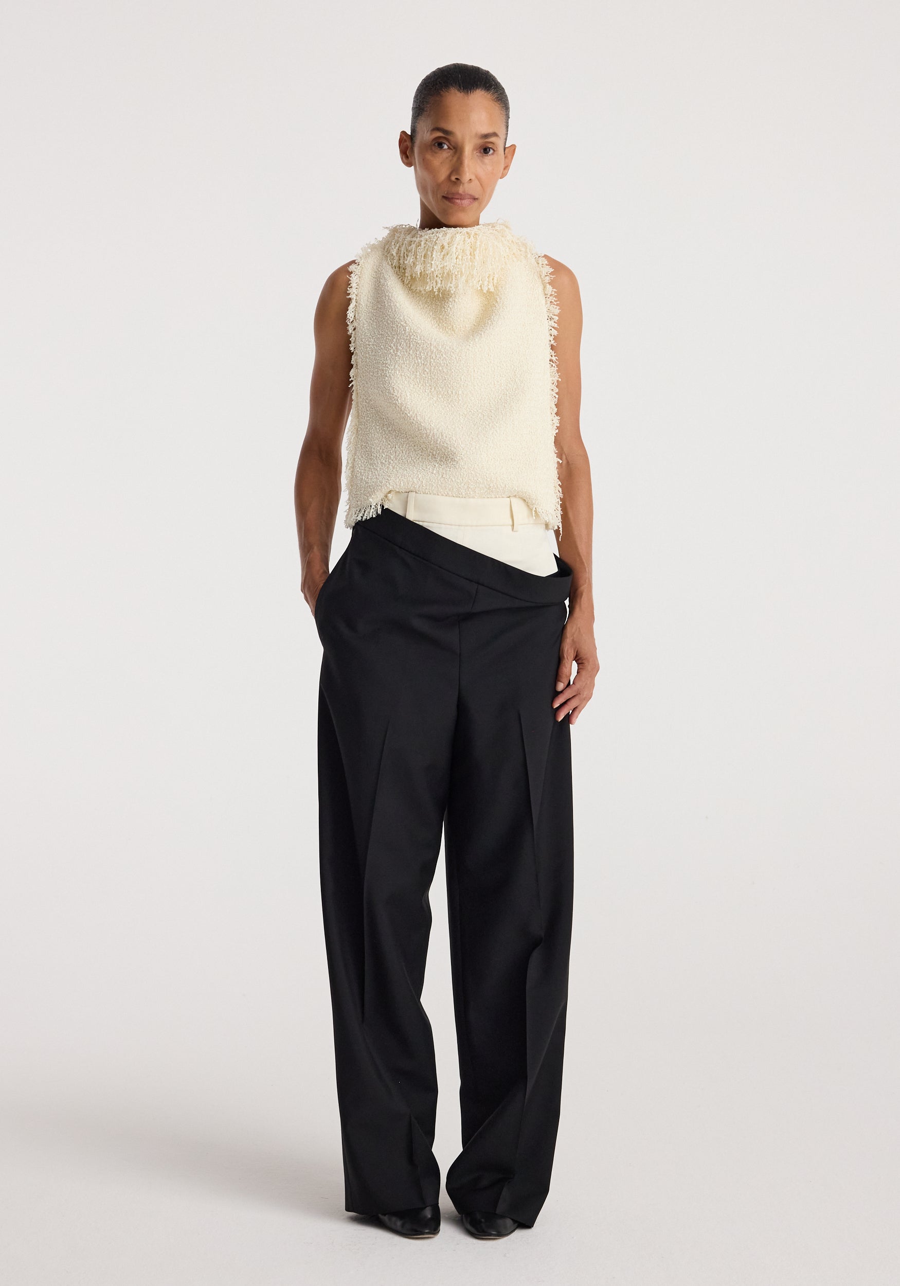 Woman wearing a sleeveless textured fringe bouclé top in cream paired with high-waisted wide-leg black trousers against a plain background.