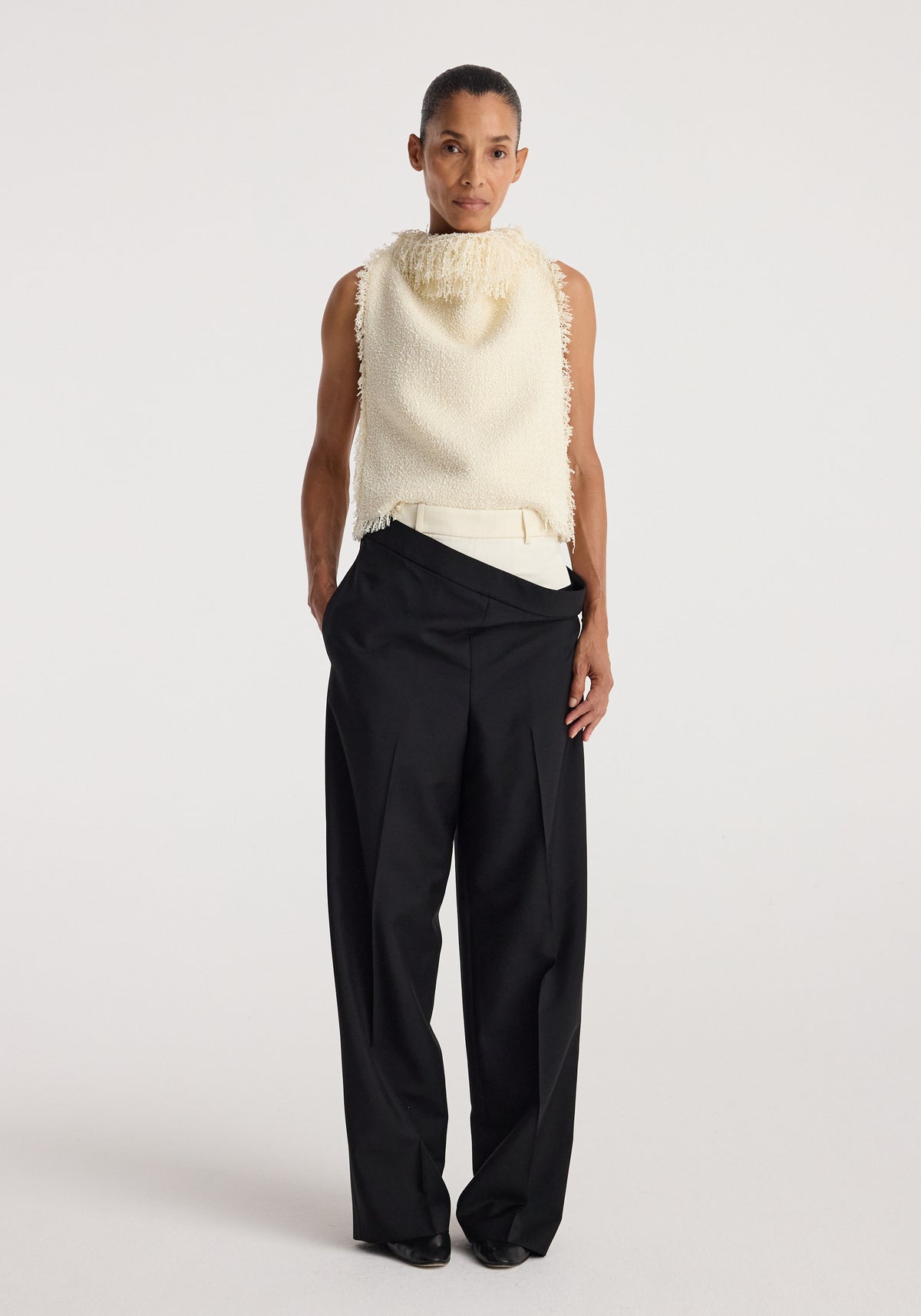 Woman wearing a sleeveless textured fringe bouclé top in cream paired with high-waisted wide-leg black trousers against a plain background.