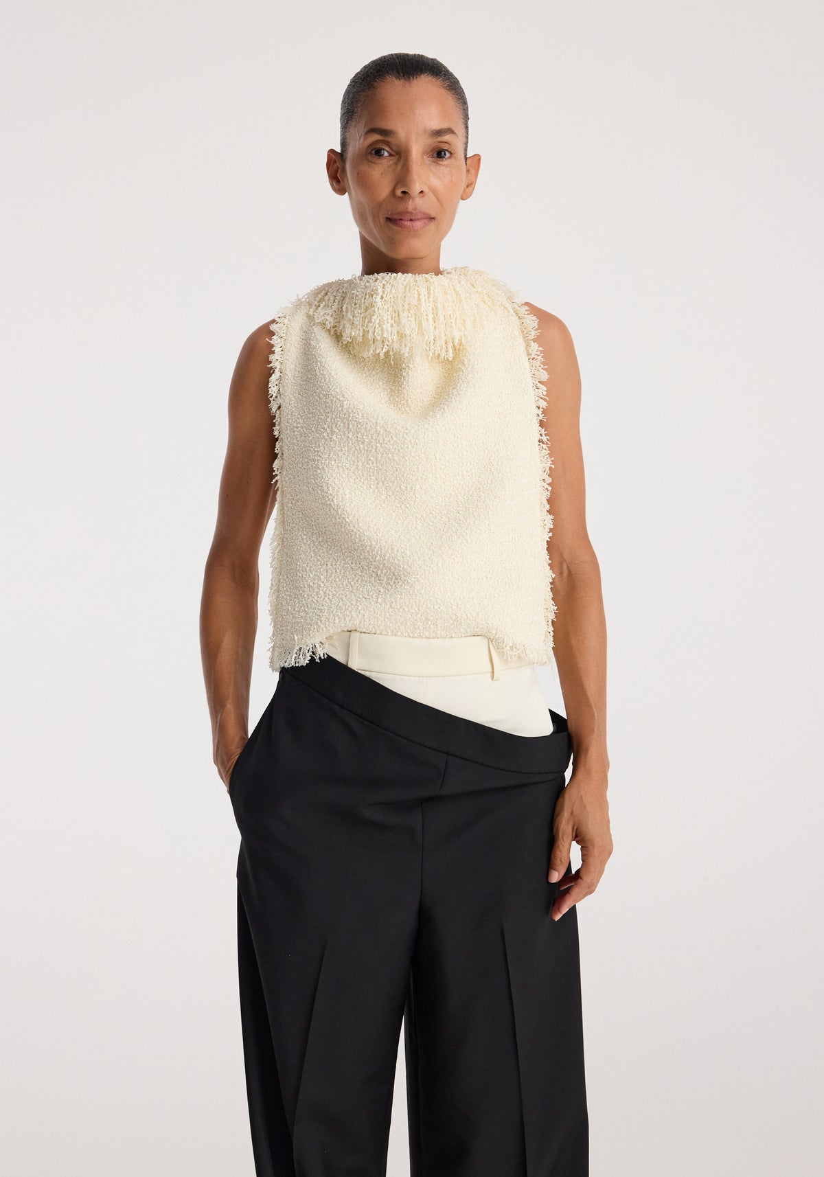 Woman wearing a sleeveless cream textured fringe bouclé top | cream with high collar and frayed edges, paired with black high-waisted pants against a plain background.