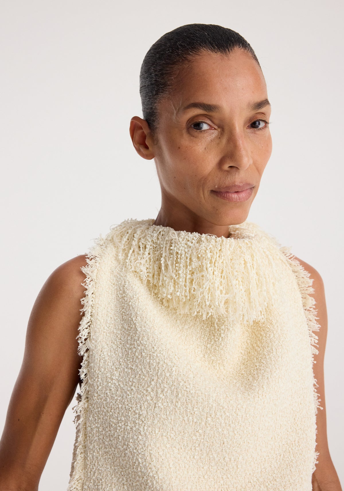 Woman wearing a sleeveless cream textured fringe bouclé top | cream with a high fringed neckline, showcasing its soft and intricate fabric details.