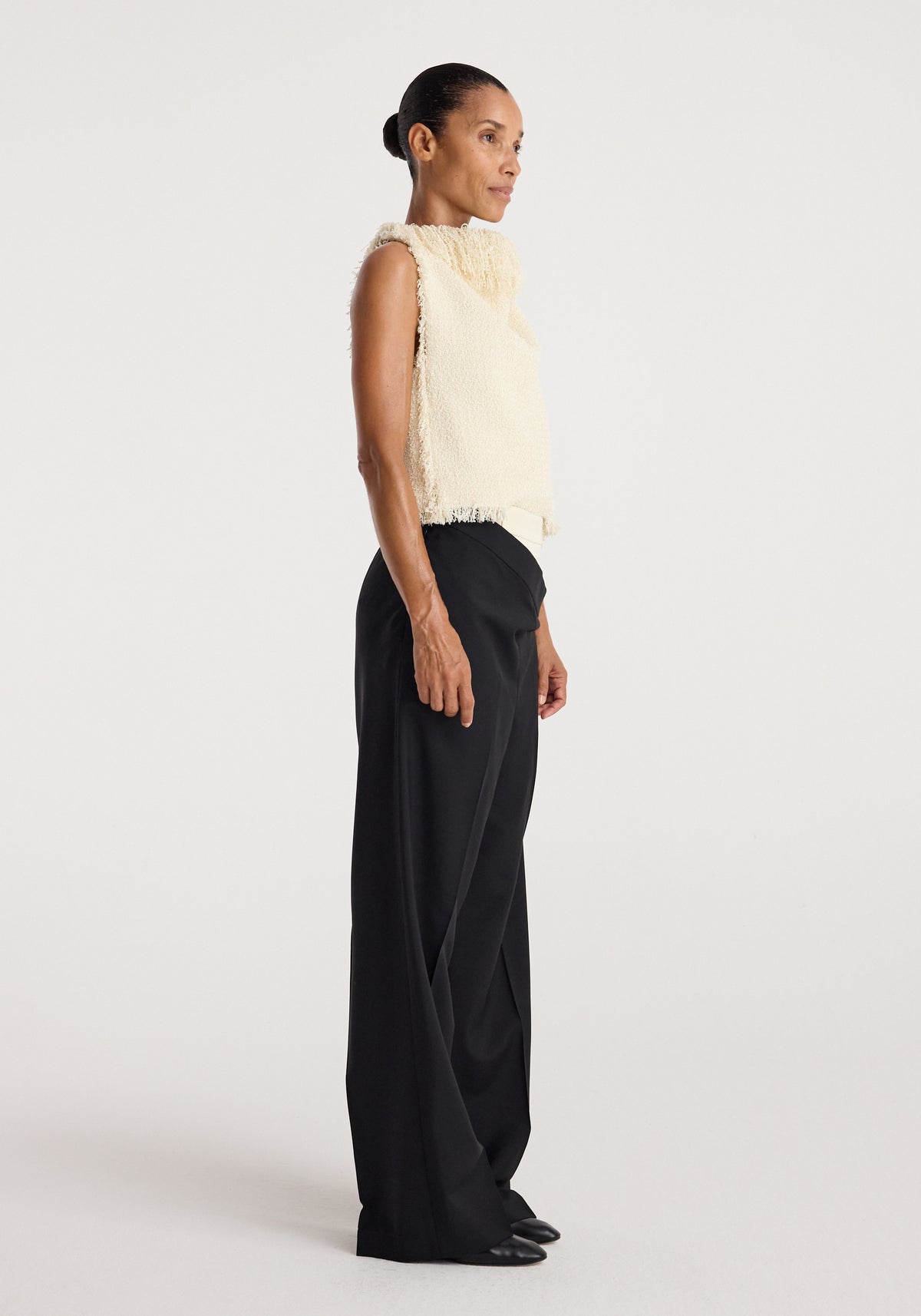 Side view of a woman wearing a sleeveless textured fringe bouclé top | cream paired with wide-leg black pants and black shoes against a plain background.