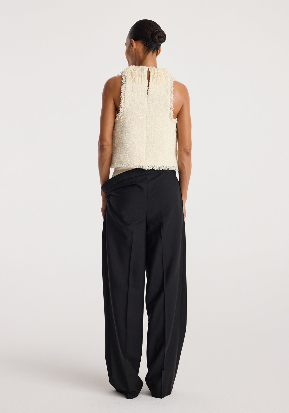 Woman seen from behind wearing a cream sleeveless textured fringe bouclé top with a keyhole back, paired with loose black wide-leg trousers against a plain background