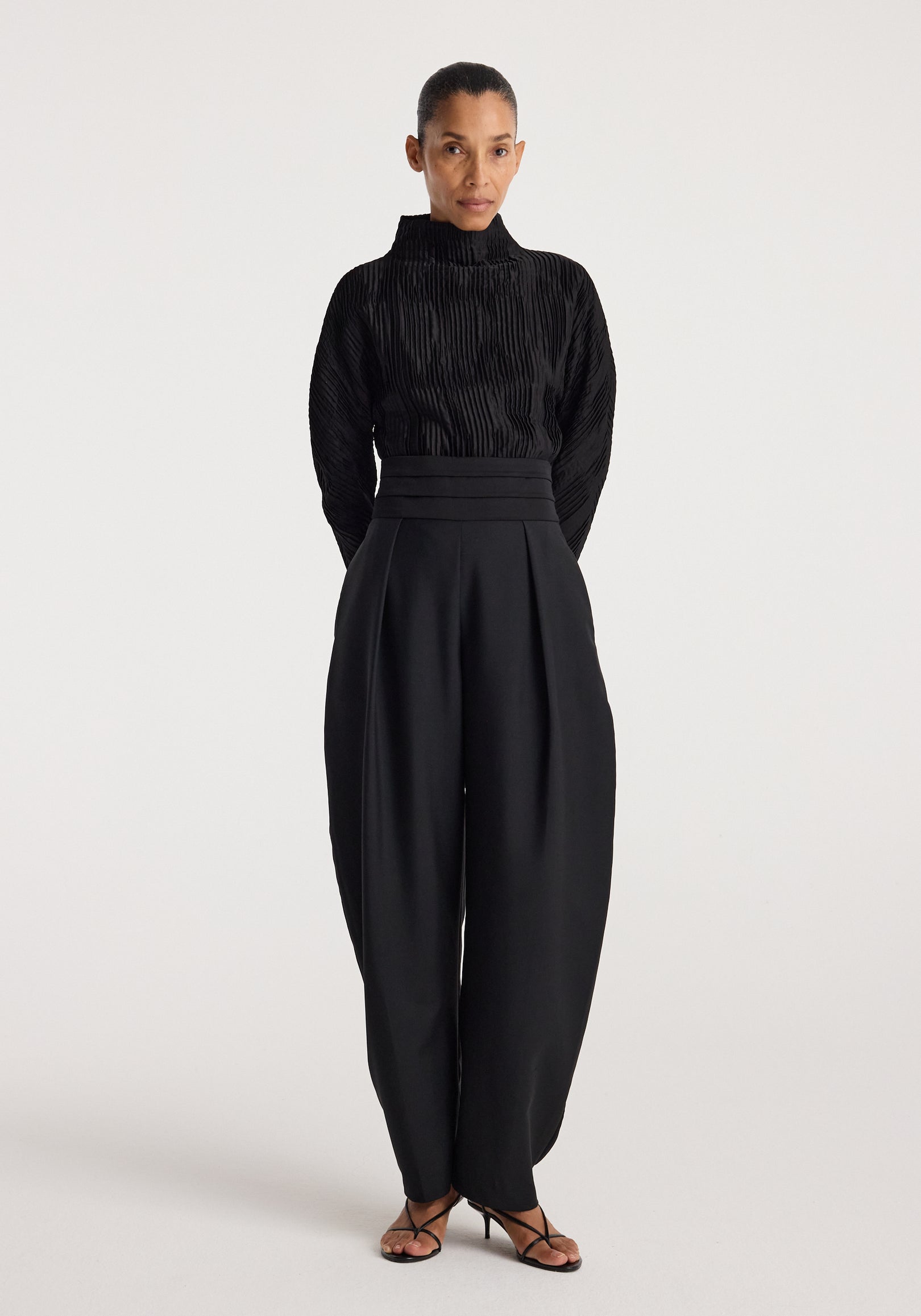 Woman wearing a black Plissé shaped top | black with long sleeves and a high neckline, paired with high-waisted wide-leg black pants and black strappy heels.