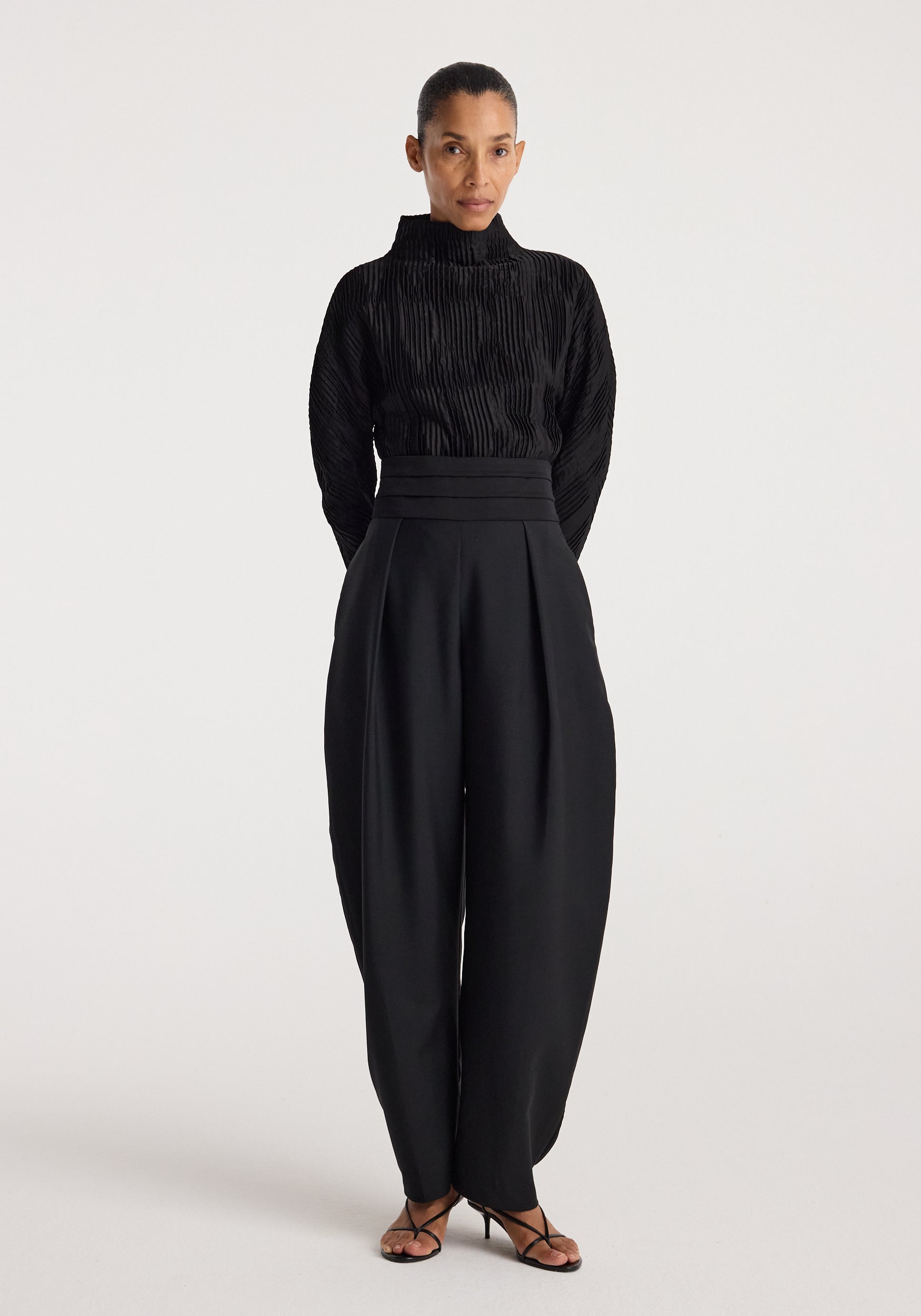 Woman wearing a black Plissé shaped top | black with long sleeves and a high neckline, paired with high-waisted wide-leg black pants and black strappy heels.