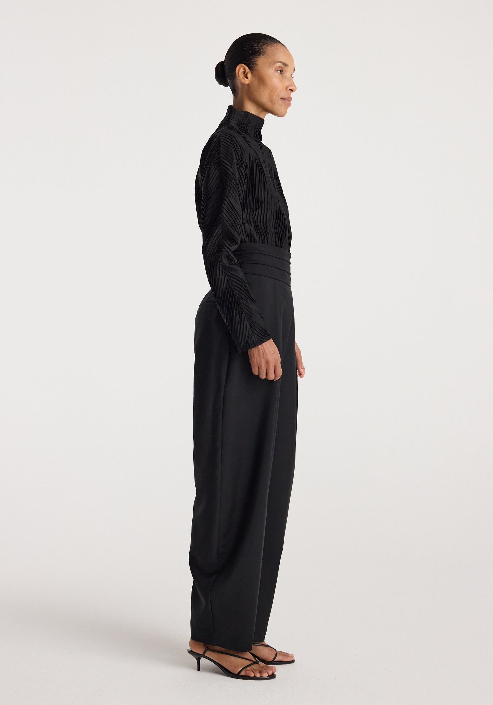 Side view of a woman wearing a long-sleeved Plissé shaped top | black with a high neckline, paired with high-waisted black pants and strappy heels.
