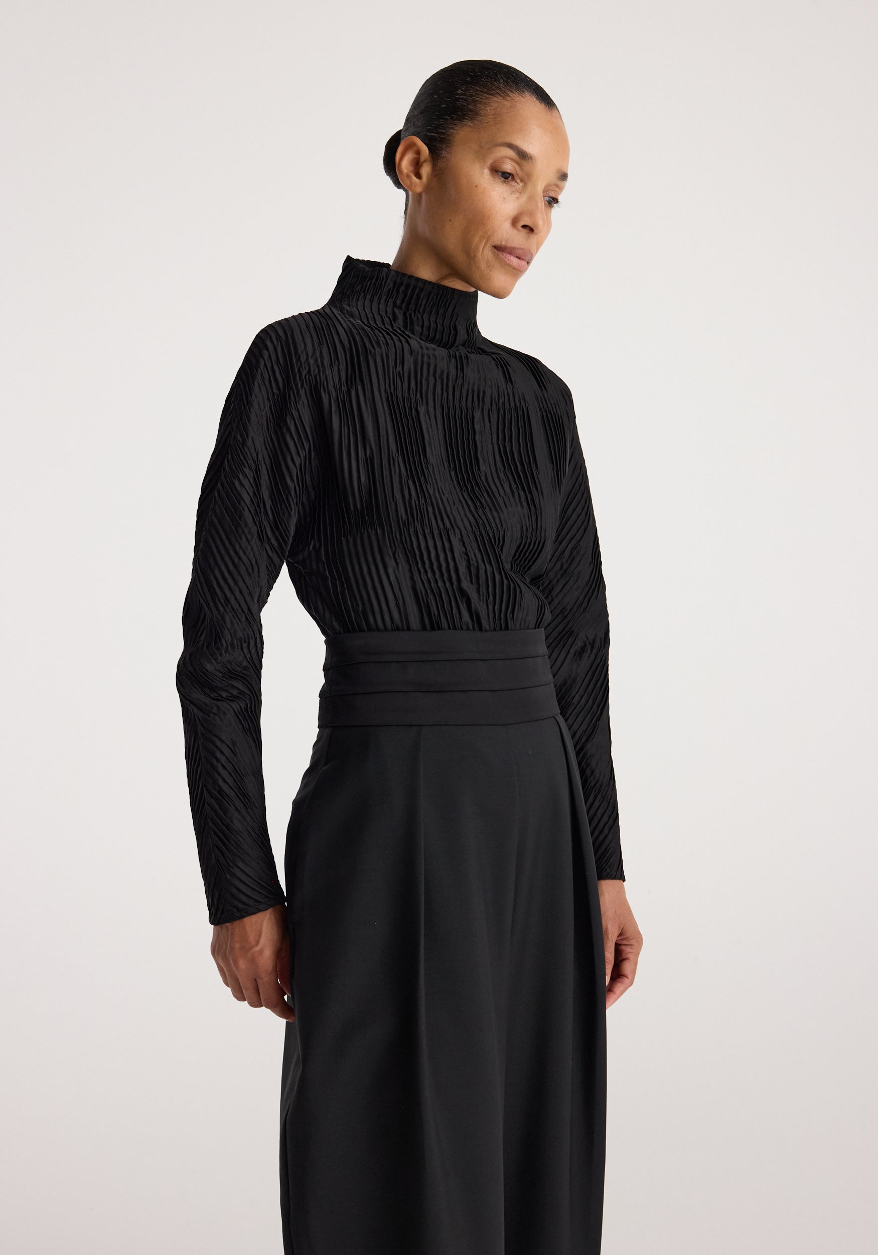 Woman wearing a black Plissé shaped top | black with a high neckline and long sleeves, styled with a high-waisted black skirt against a neutral background.