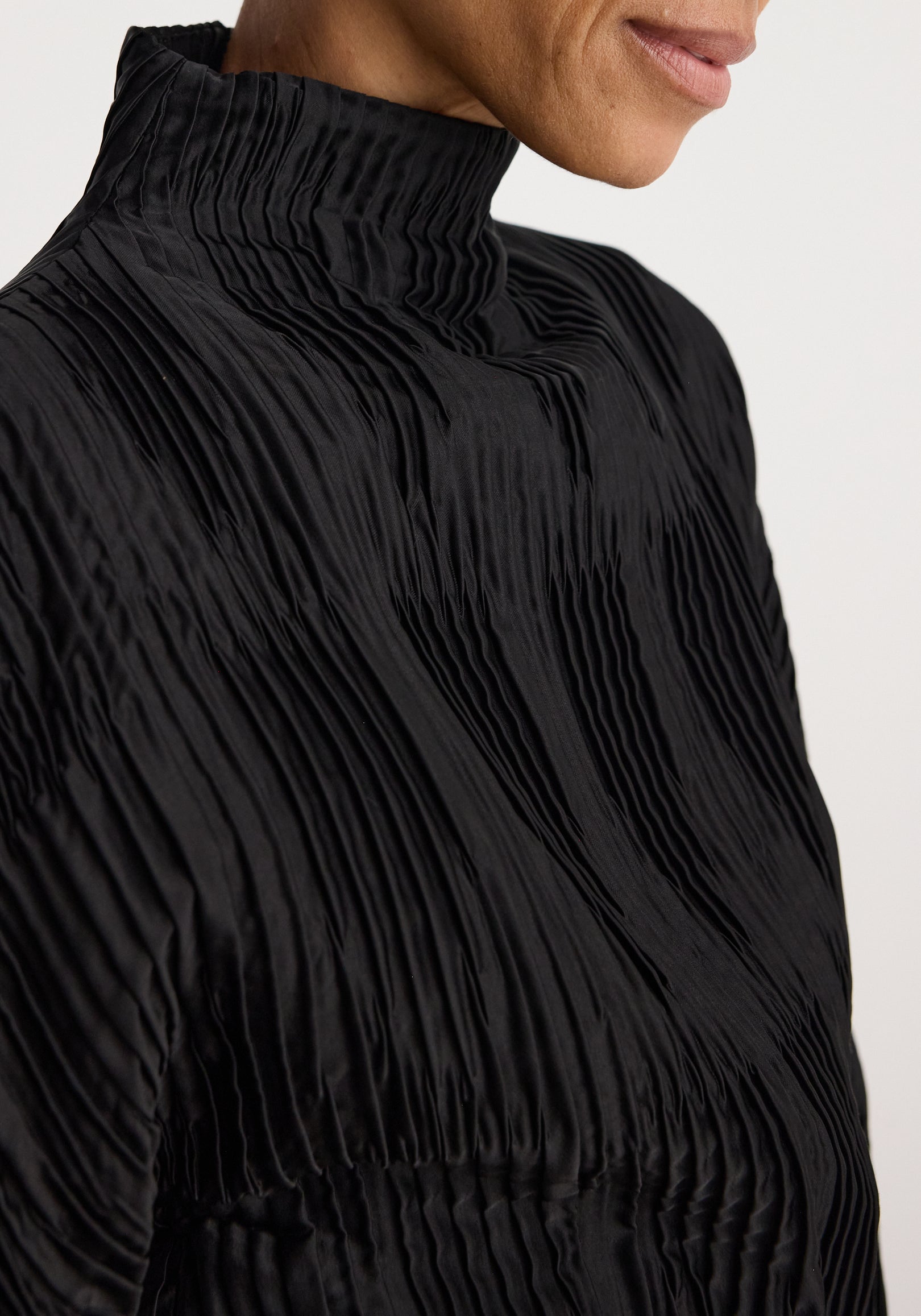 Close-up of a person wearing a Plissé shaped top | black featuring a high neck and textured pleated fabric that creates elegant vertical lines.