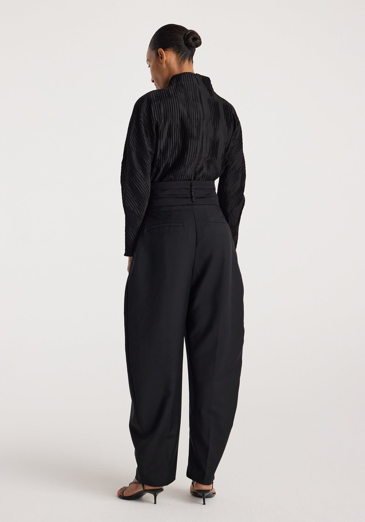 Woman wearing a Plissé shaped top | black with long sleeves and a high neckline, paired with black high-waisted pants and black heeled sandals, standing against a plain background.