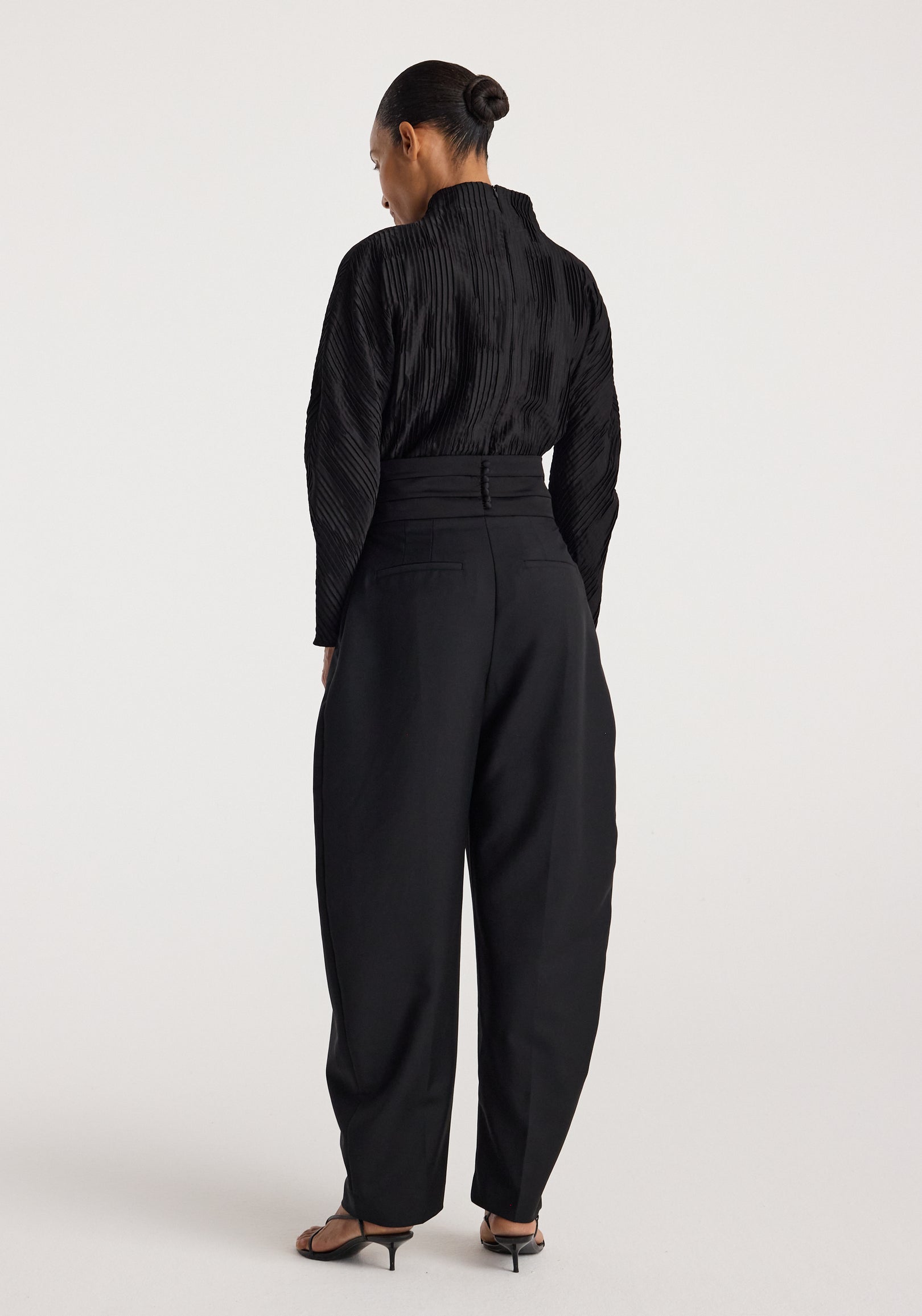 Woman wearing a Plissé shaped top | black with long sleeves and a high neckline, paired with black high-waisted pants and black heeled sandals, standing against a plain background.