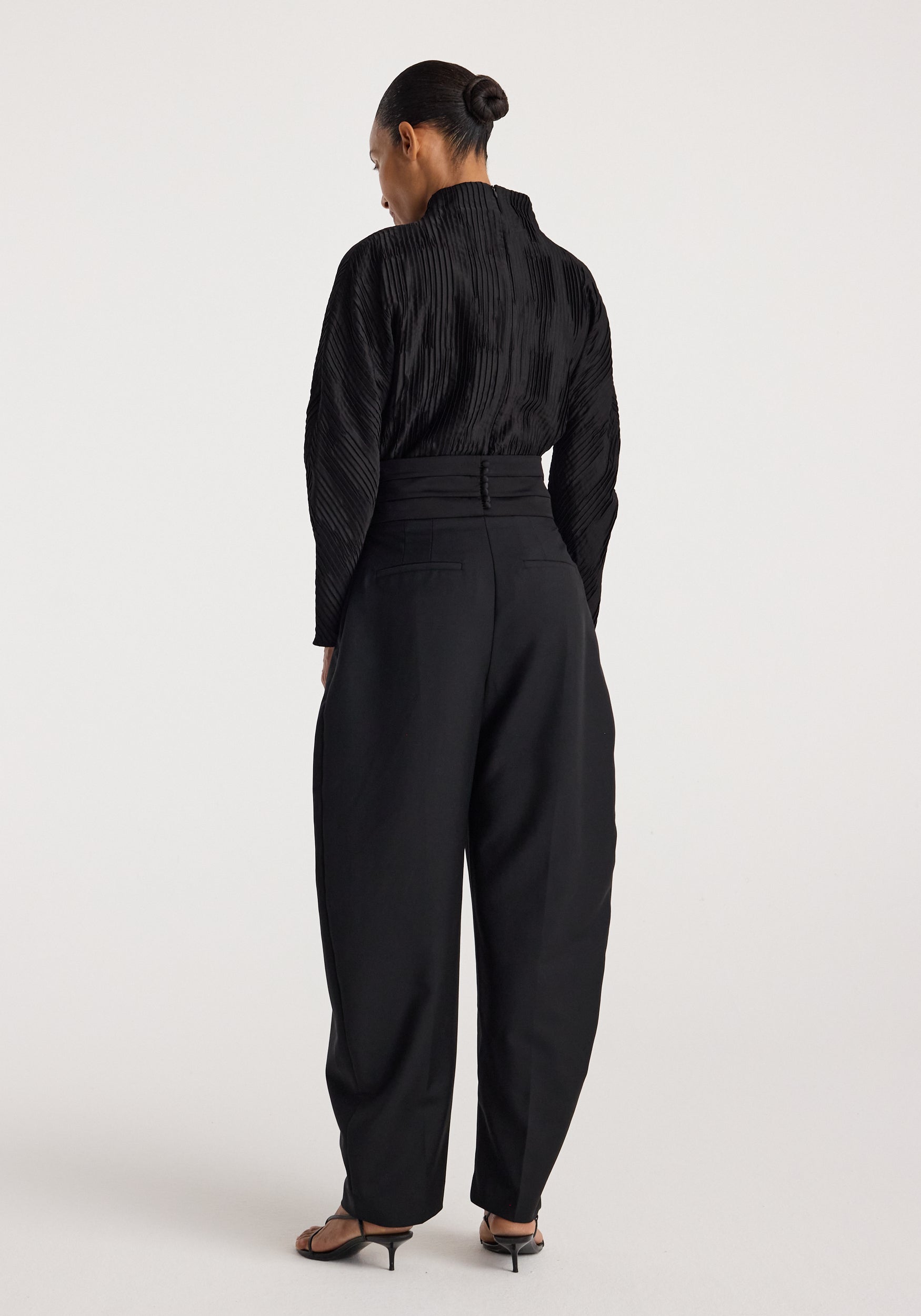 Woman wearing a Plissé shaped top | black with long sleeves and a high neckline, paired with black high-waisted pants and black heeled sandals, standing against a plain background.