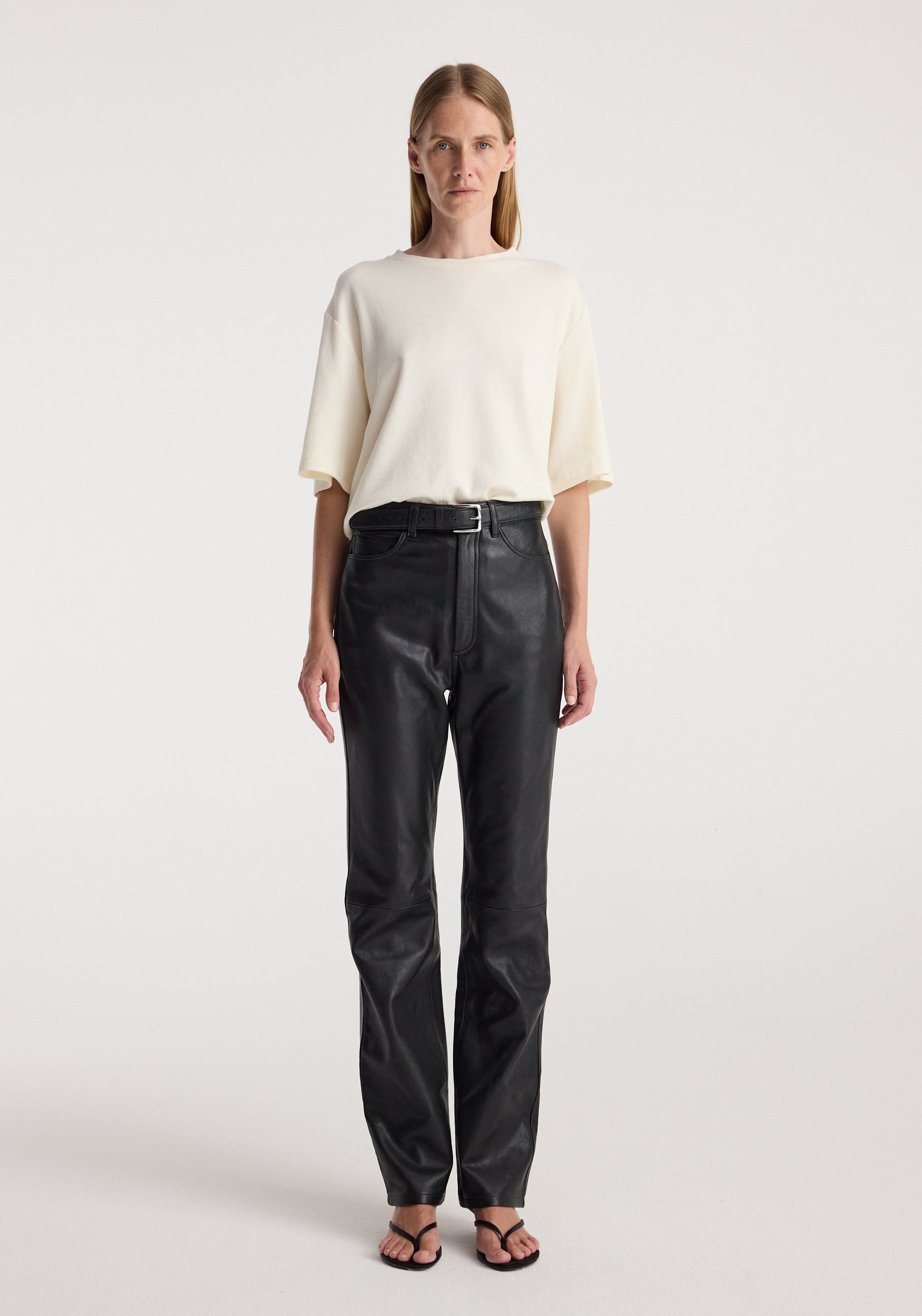 Woman wearing an oversized cotton wool t-shirt | cream paired with high-waisted black leather pants and black open-toe sandals against a plain background.